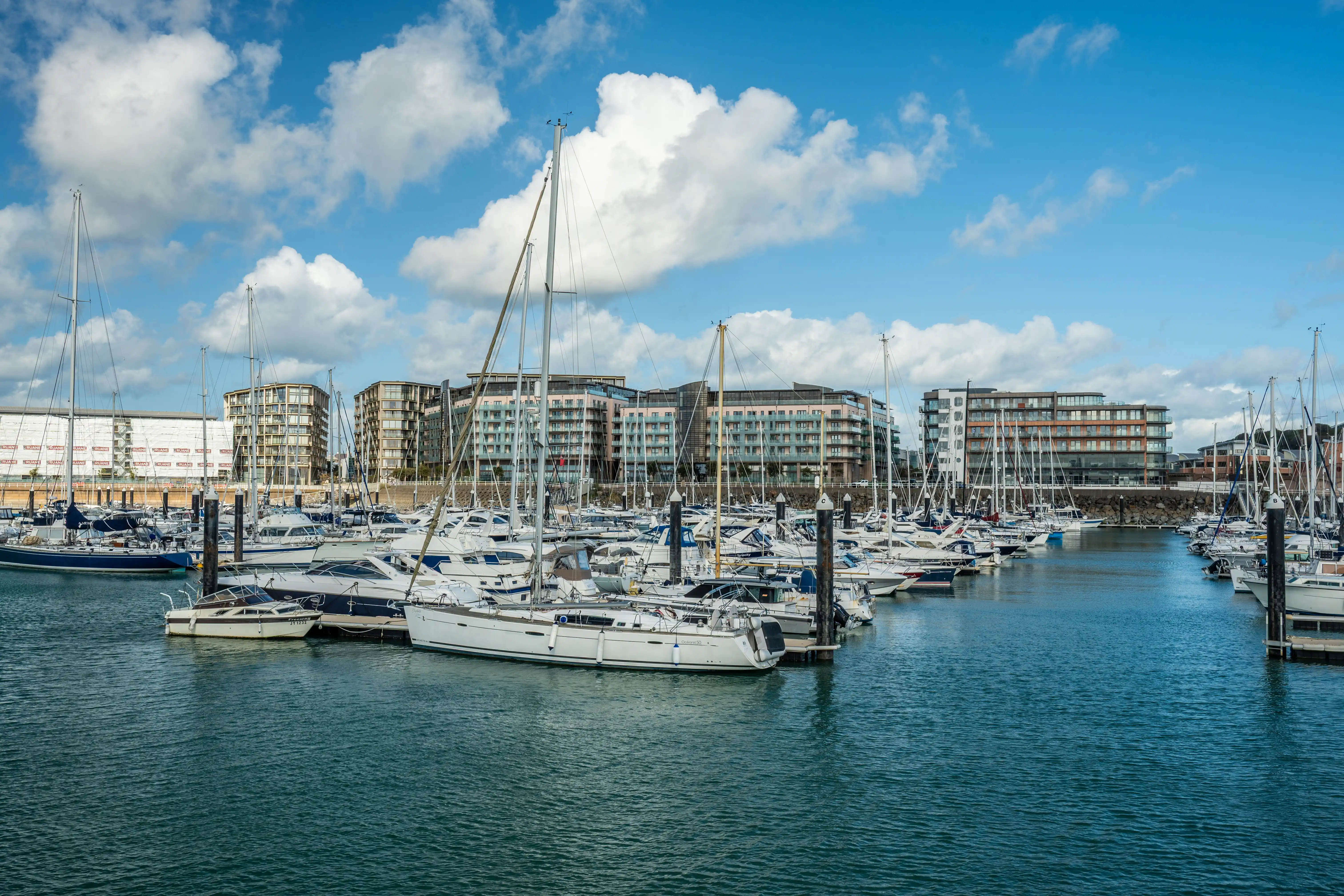 Numerous white sailing yachts and motorboats are docked in a blue harbour. In the background, modern, multi-storey apartment buildings with large windows line the waterfront. The sky is blue with scattered white clouds.