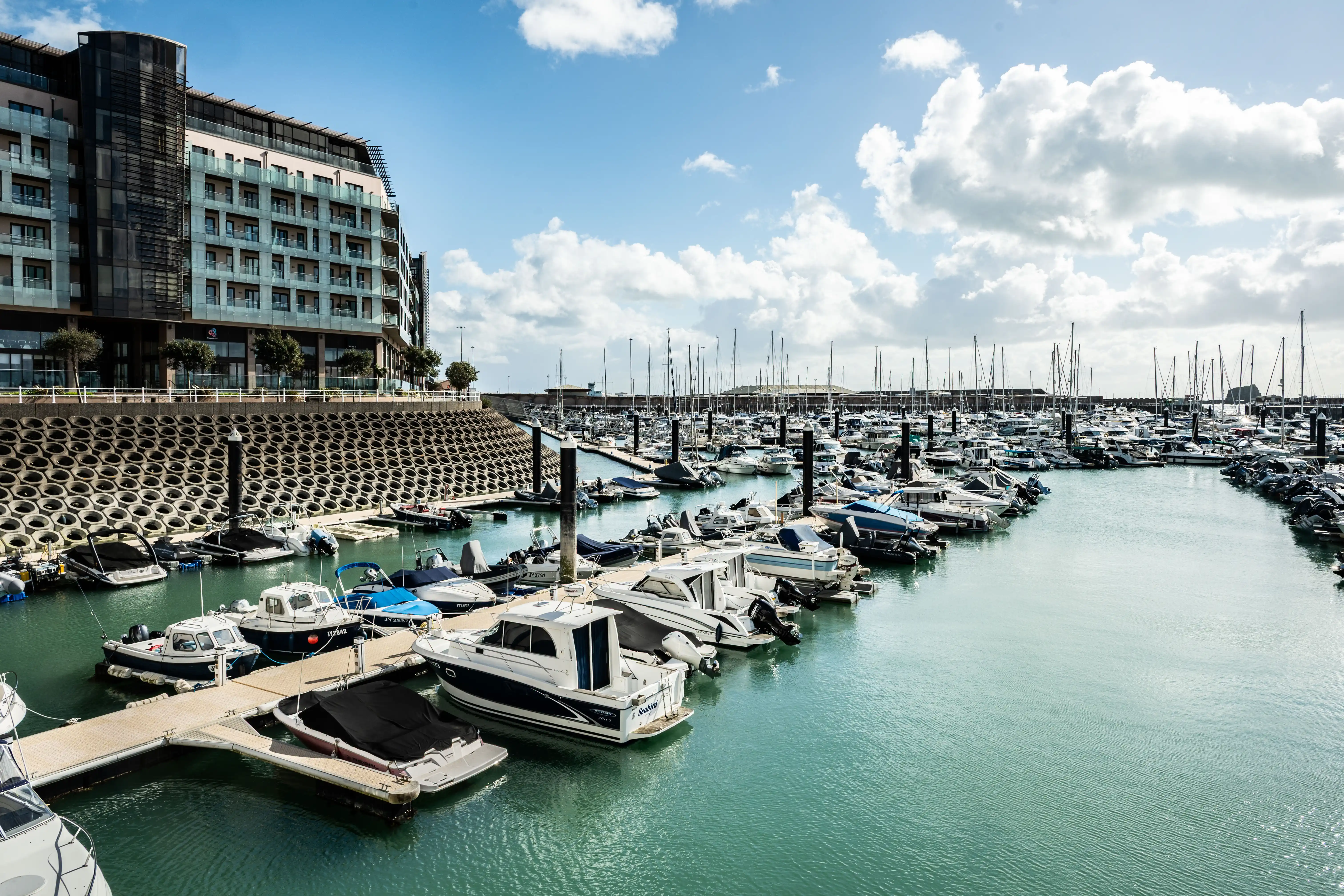 A marina harbour with numerous boats and yachts docked, a modern multi-story building to the left, and a textured concrete sea wall.
