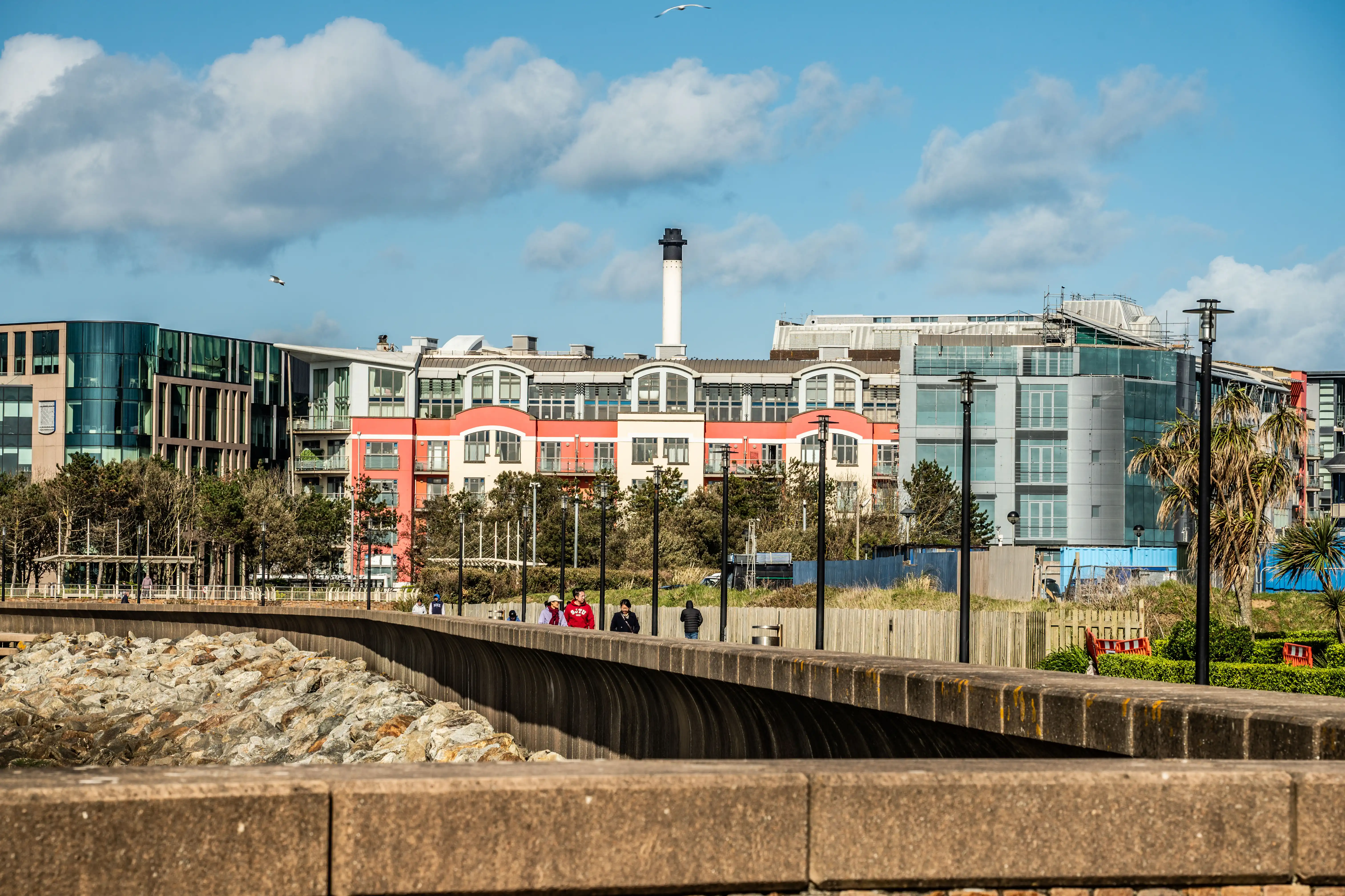 A modern seaside promenade with contemporary buildings in the background, featuring a red and cream coloured block of apartments. People are walking along the path beside a sea wall made of large stones.