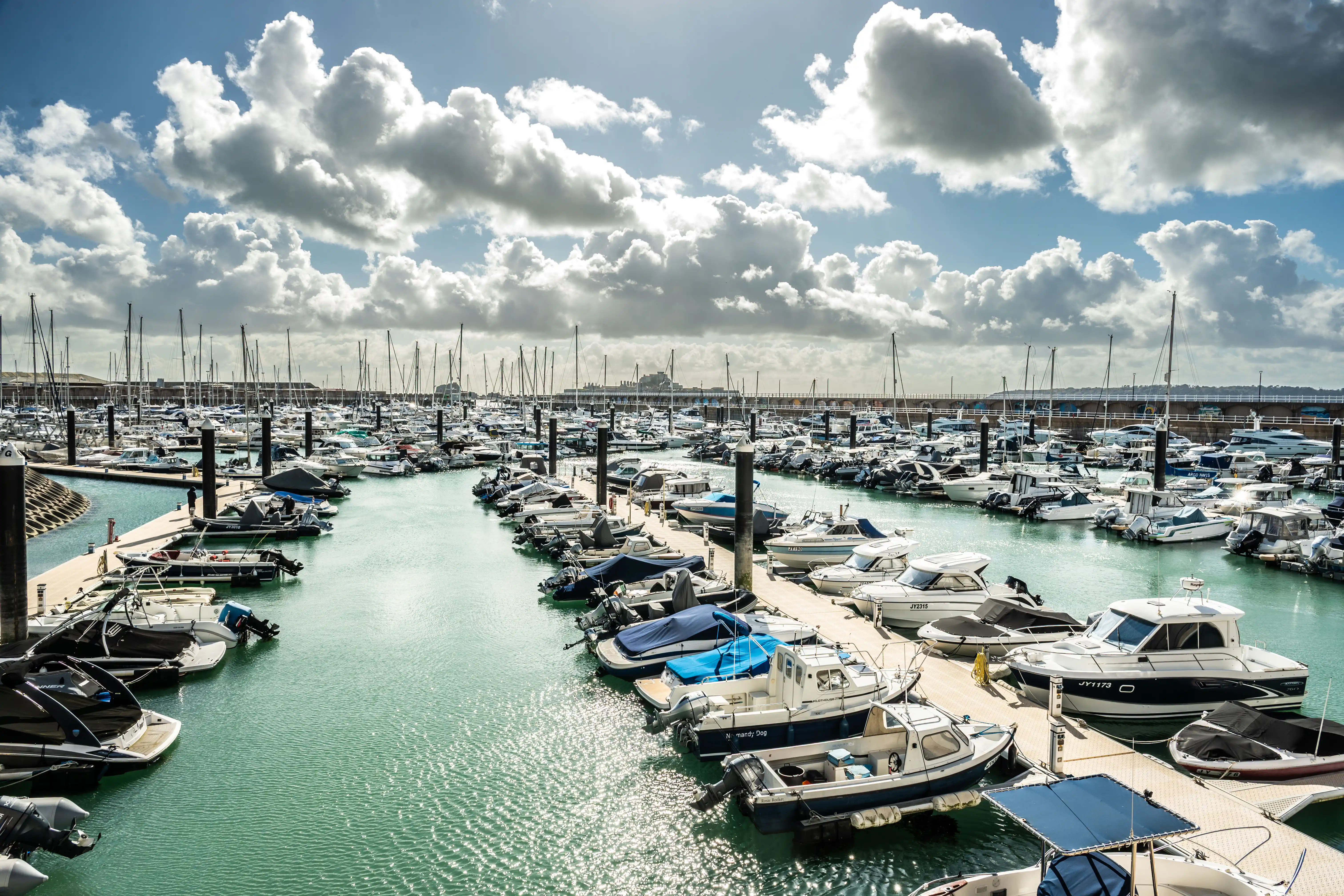 A panoramic view of a crowded marina with many boats docked. The water is a vibrant turquoise, and the sky is filled with large, white, fluffy clouds interspersed with patches of blue. Sunlight streams through the clouds, creating highlights on the water and boats.