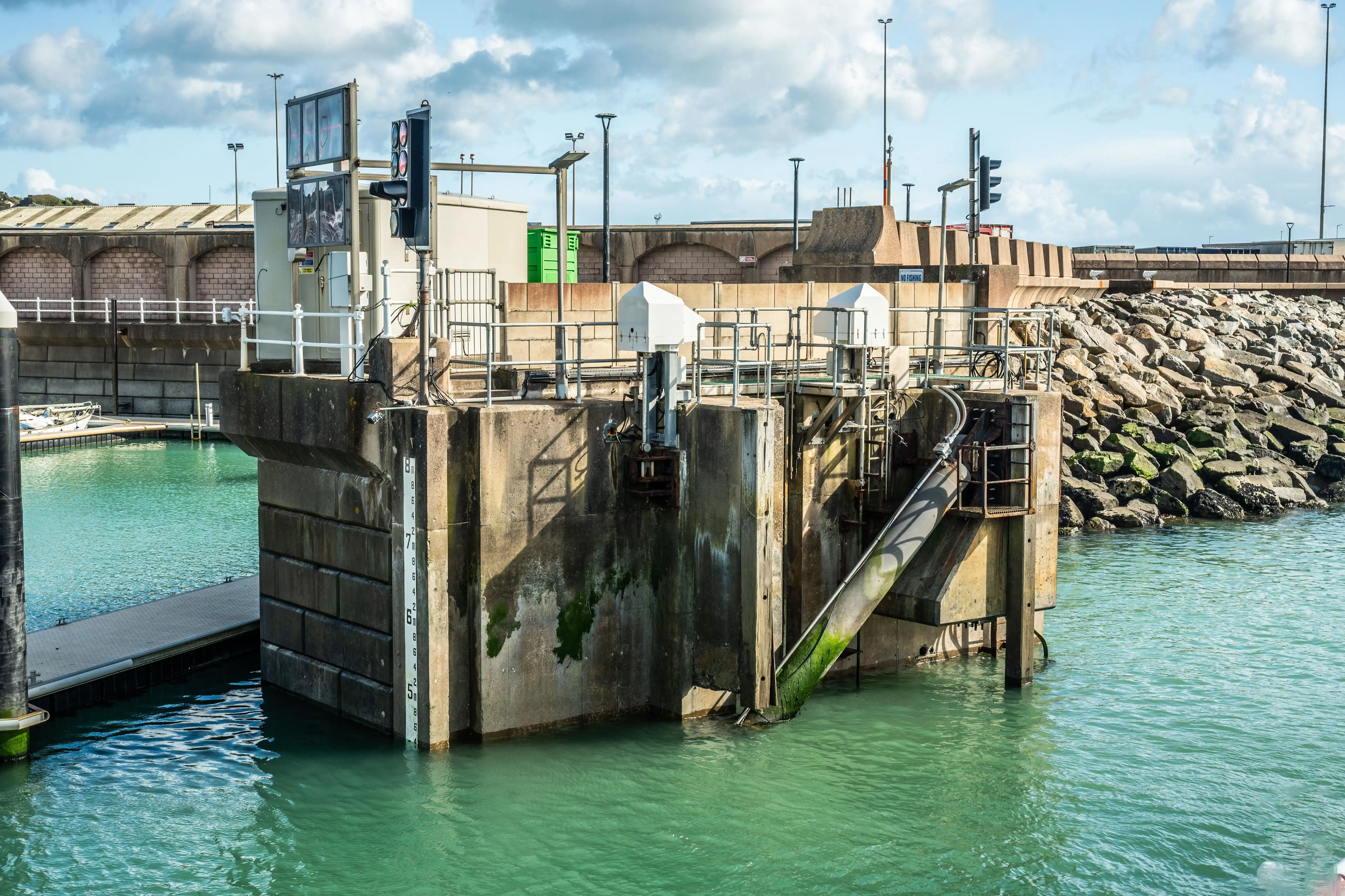 A sturdy concrete maritime structure stands in clear turquoise water, featuring a visible water level gauge on its side and traffic lights for vessels. Rocky breakwaters and harbour buildings are visible in the background under a partly cloudy sky.