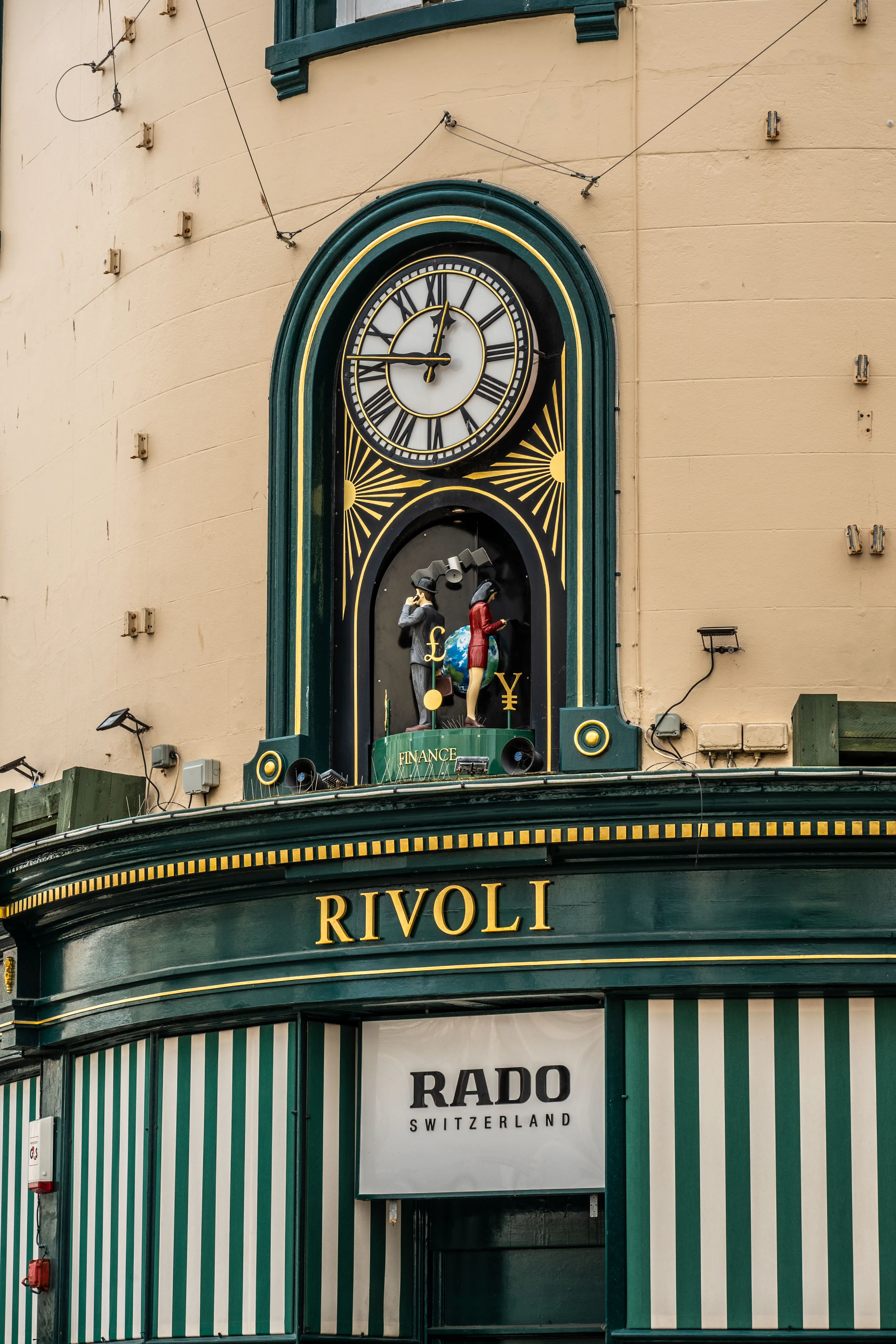 Close-up of a building facade featuring an arched clock with Roman numerals, a kinetic sculpture symbolising finance, the word RIVOLI in gold lettering, and a RADO SWITZERLAND sign with green and white striped awnings.