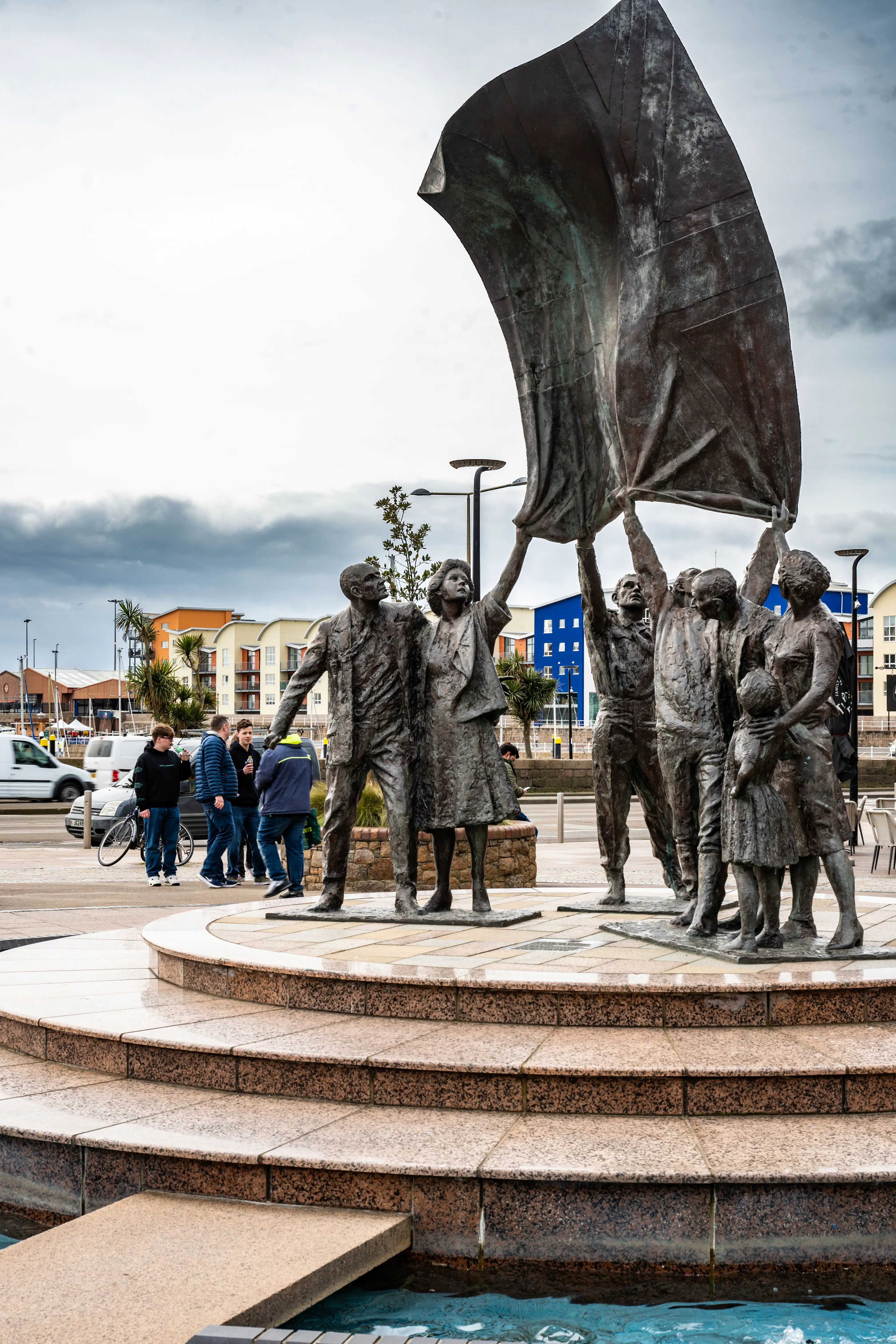 Bronze sculpture of a group of people hoisting a large, unfurled flag, standing on a tiered granite pedestal with water at its base. Modern buildings and a cloudy sky are visible in the background.
