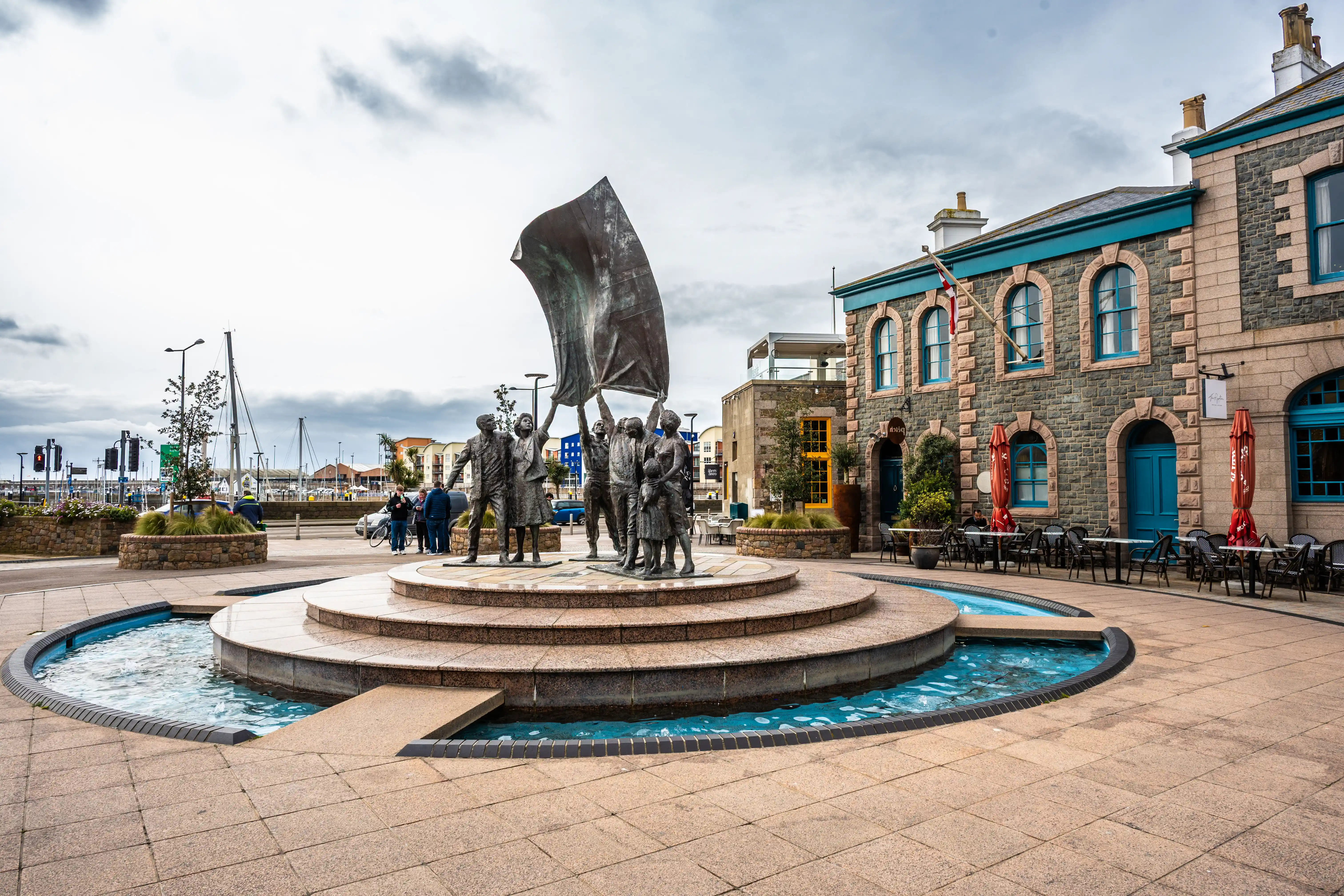 A bronze sculpture of several people holding up a large flag stands in the centre of a circular fountain in a paved square. Behind the sculpture is a stone building with blue windows and doors, with outdoor cafe seating.