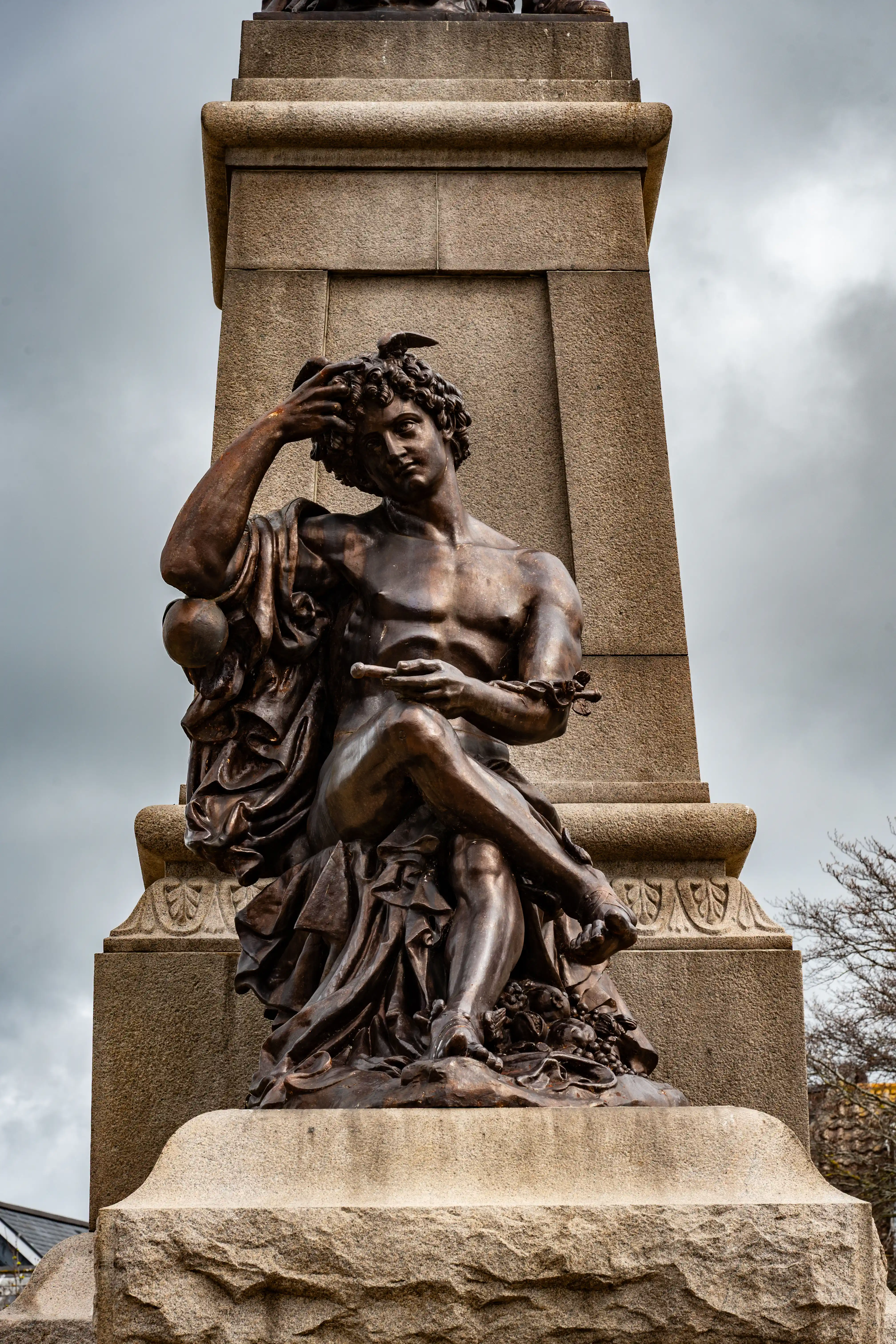 A dark bronze statue of a man with curly hair, sitting with his arm raised to his head, is placed against a large stone monument. A small bird is visible on his head.