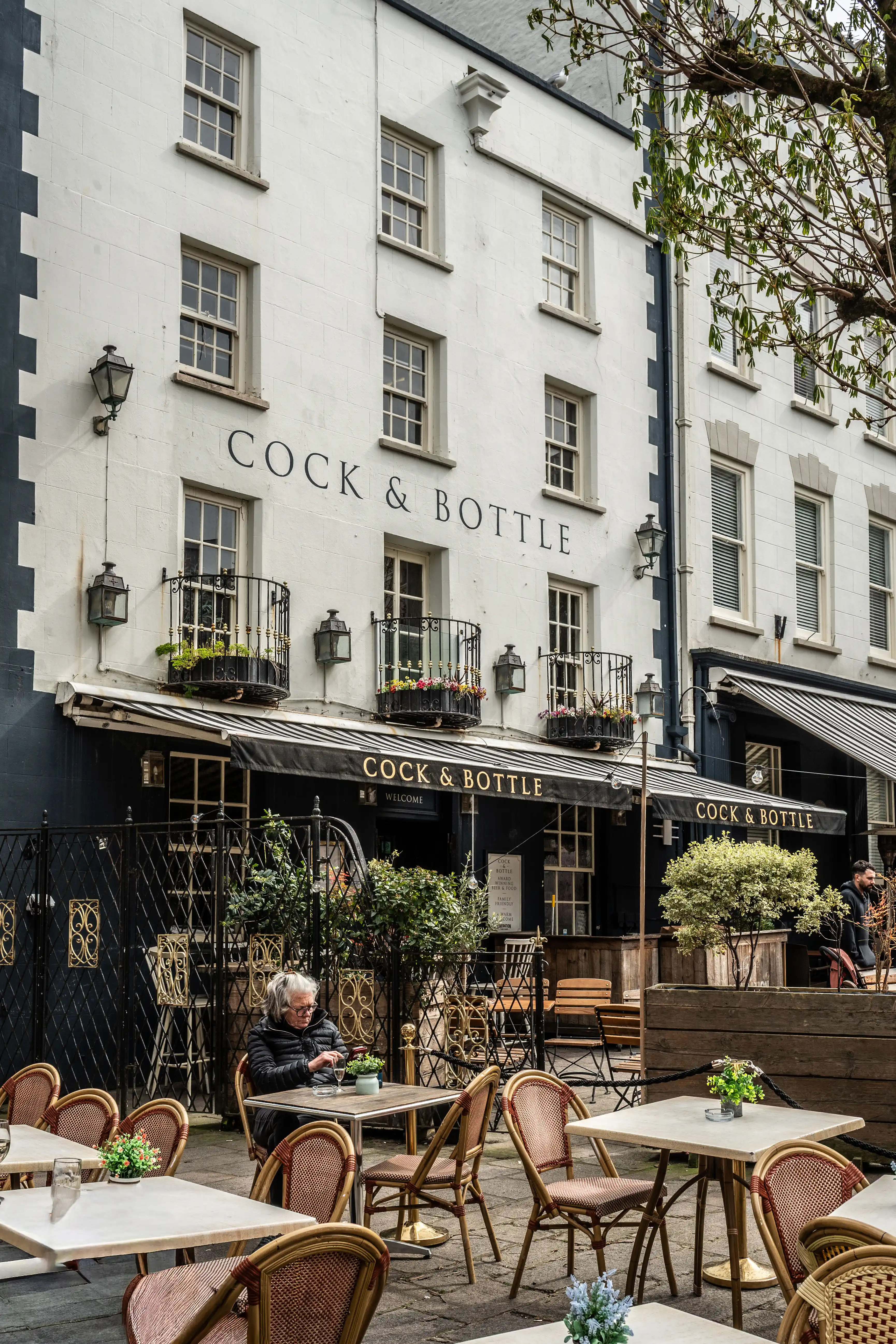 A street-level view of the Cock & Bottle pub, showcasing its traditional architecture and outdoor seating area. An elderly woman with grey hair, wearing a dark jacket, is seated at a small table with a drink, engrossed in her mobile phone.