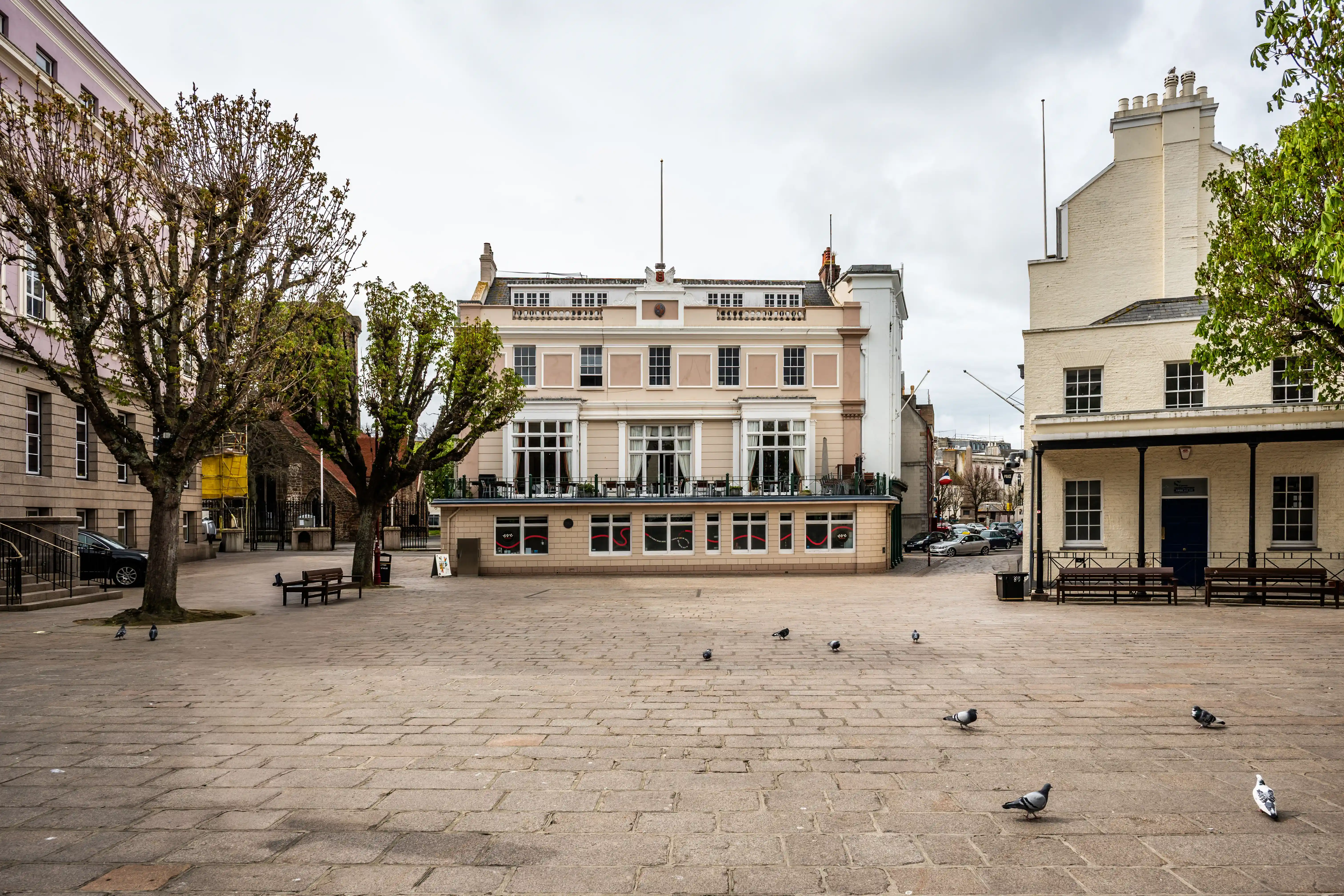 This photograph captures a wide, open paved square. In the background, a large, pale pink building with numerous windows dominates the scene. It has a classical architectural style with ornate detailing at the top. To the right of this building is a smaller white building. Several trees are scattered around the square, and a few pigeons are pecking at the ground. A lone woman is walking away from the camera.