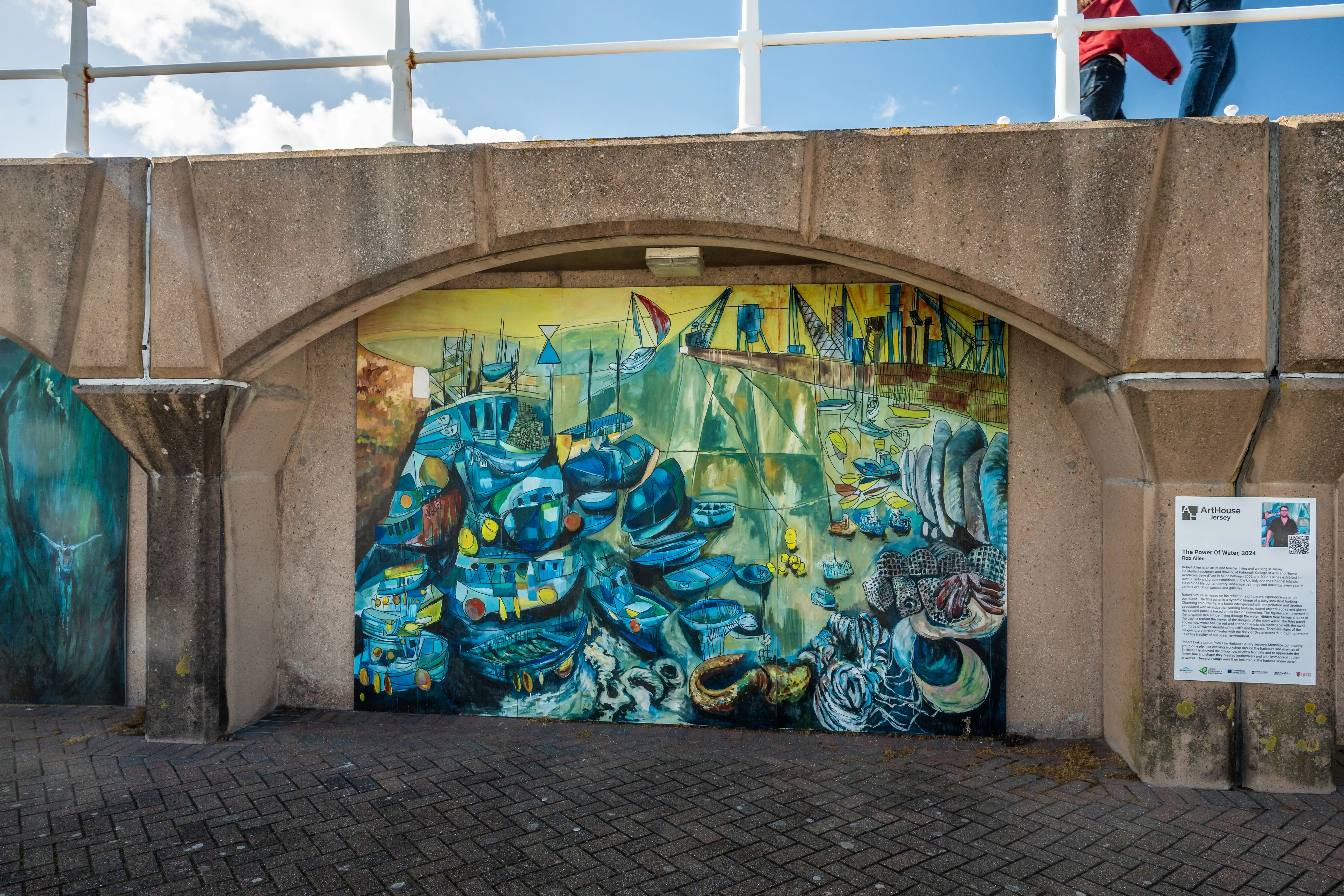 A bright, detailed mural of a crowded harbour with many blue boats, cranes, and industrial elements, painted on a wall under a concrete archway. Another mural is visible to the left, and an information board is on the right. The scene is set against a backdrop of a blue sky with clouds and a paved walkway.