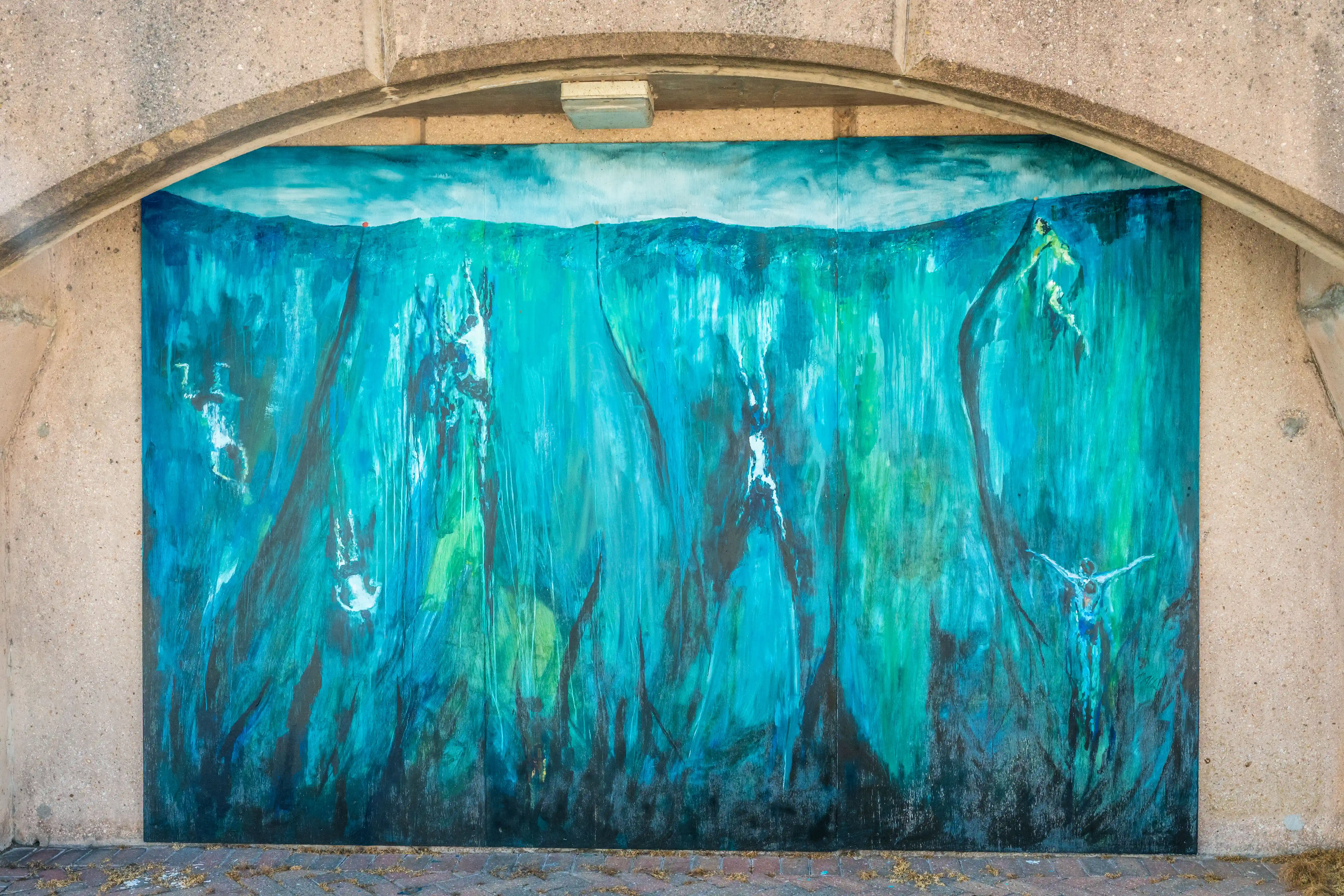 Abstract mural of an underwater scene with multiple figures in shades of blue and green, mounted on a wall under a concrete archway.