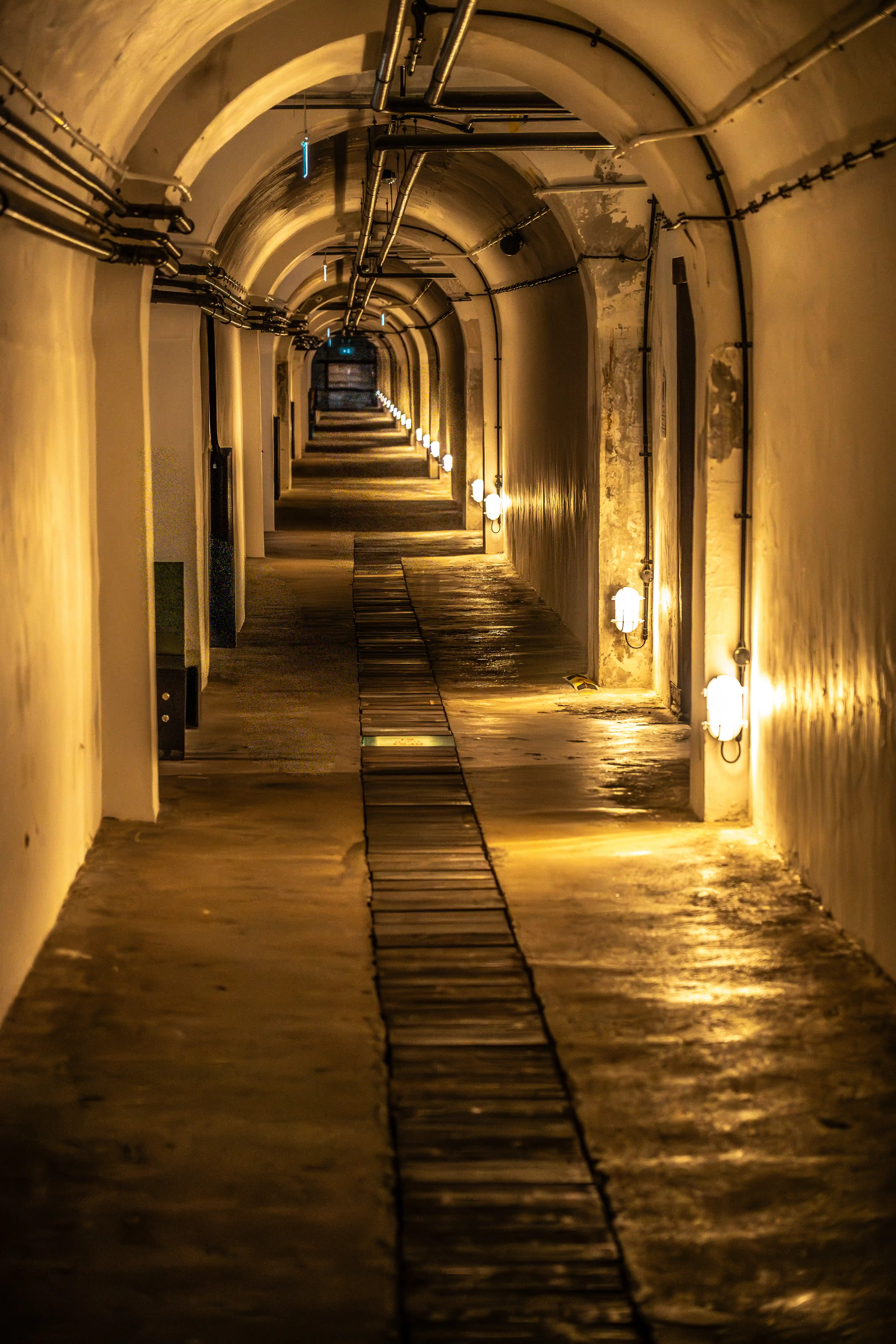 Interior view of a long, curved tunnel with white arched ceilings. Industrial piping runs along the top of the walls. Numerous wall-mounted light fixtures emit a warm, yellow light, reflecting off the wet, concrete floor. A wooden plank walkway is laid down the middle of the tunnel.