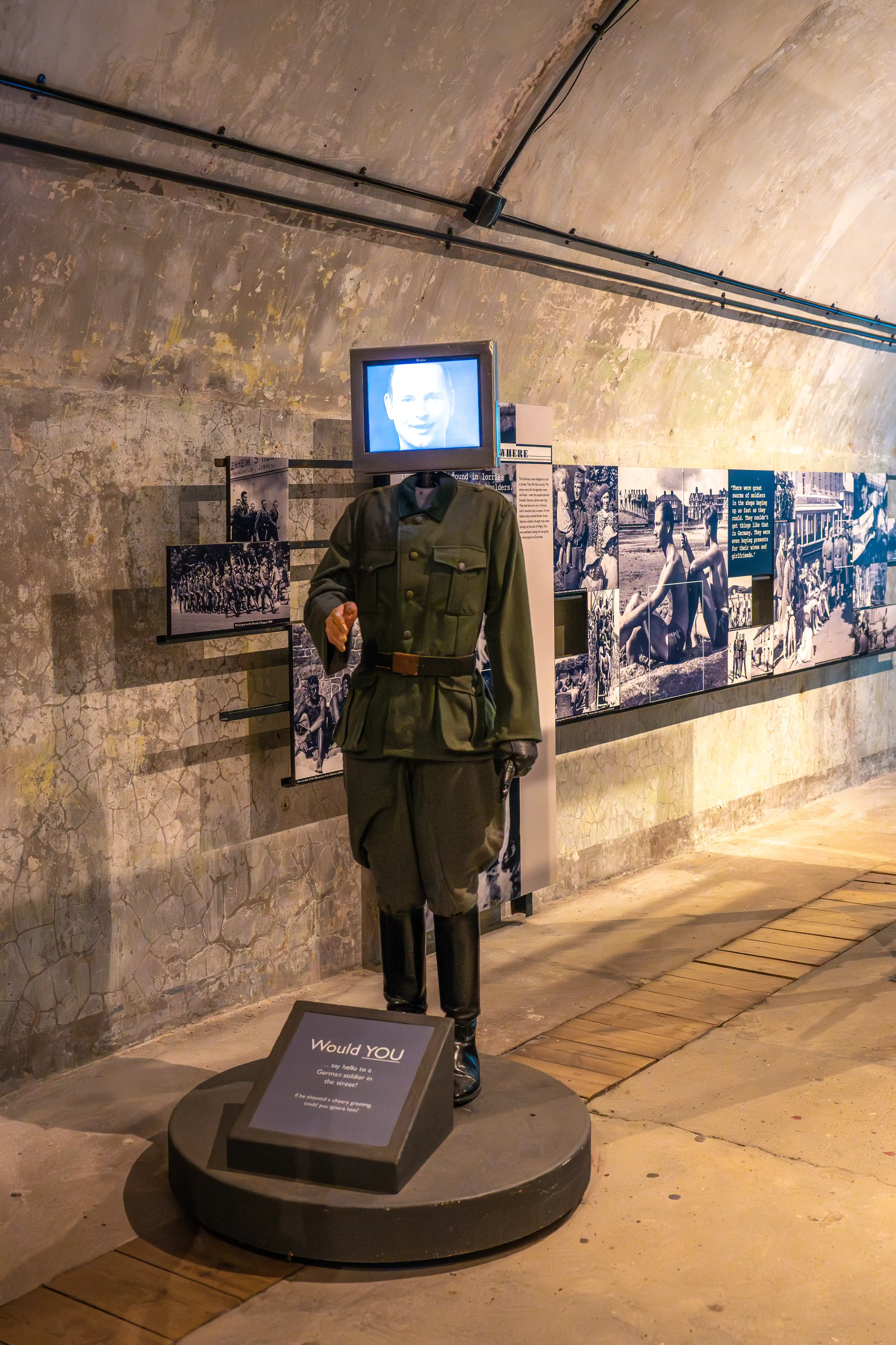Mannequin in a German WWII uniform with a television screen displaying a portrait of a man as its head, stands in a museum exhibit.