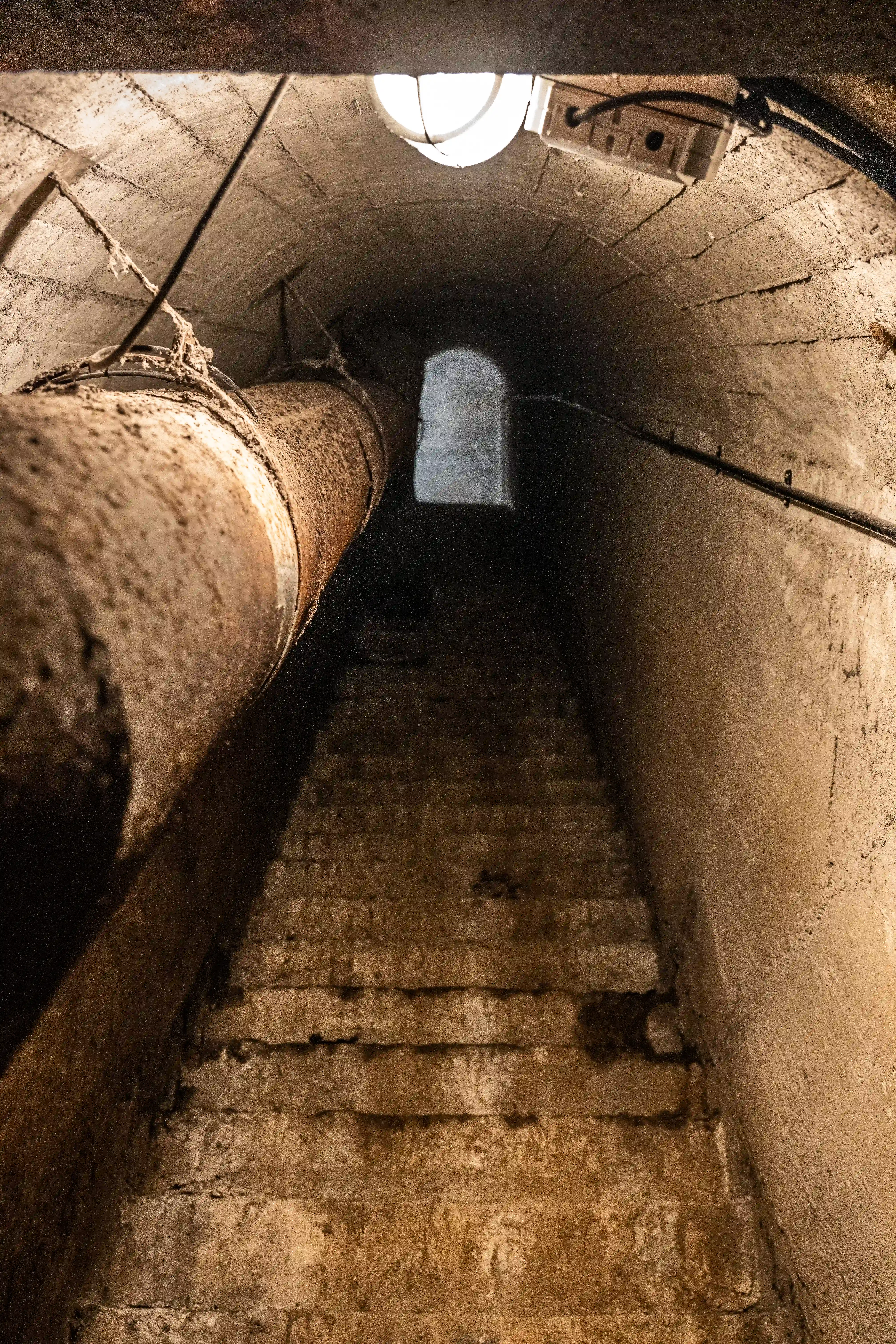 A rusty pipe runs along the left side of a dark, concrete tunnel with worn stairs leading downwards. A light fixture is visible at the top, and an arched opening is at the end of the tunnel.