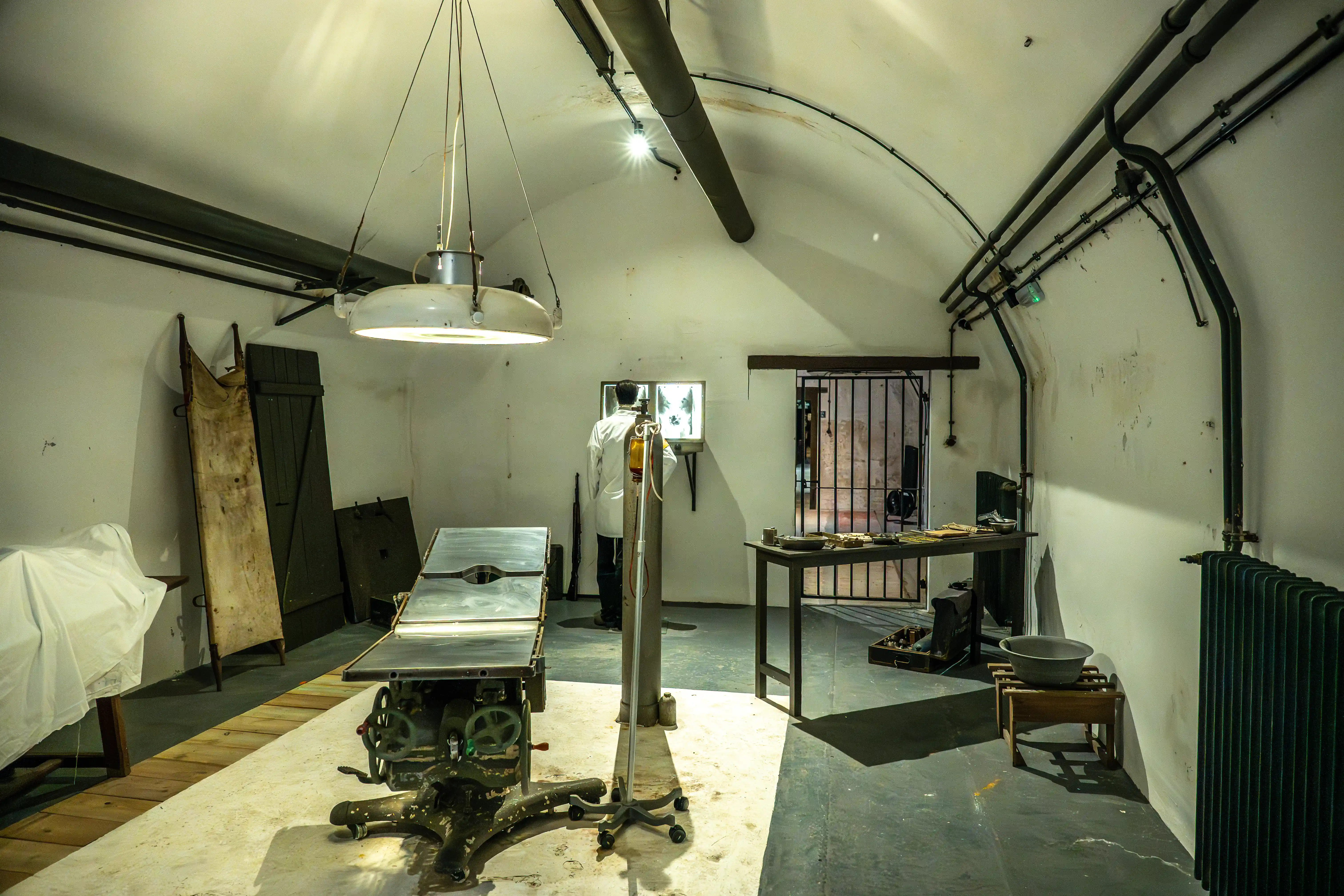  Interior of a vintage operating theatre in a bunker, featuring a medical examination table, a stretcher, a doctor in a white coat looking at X-rays, and exposed pipework