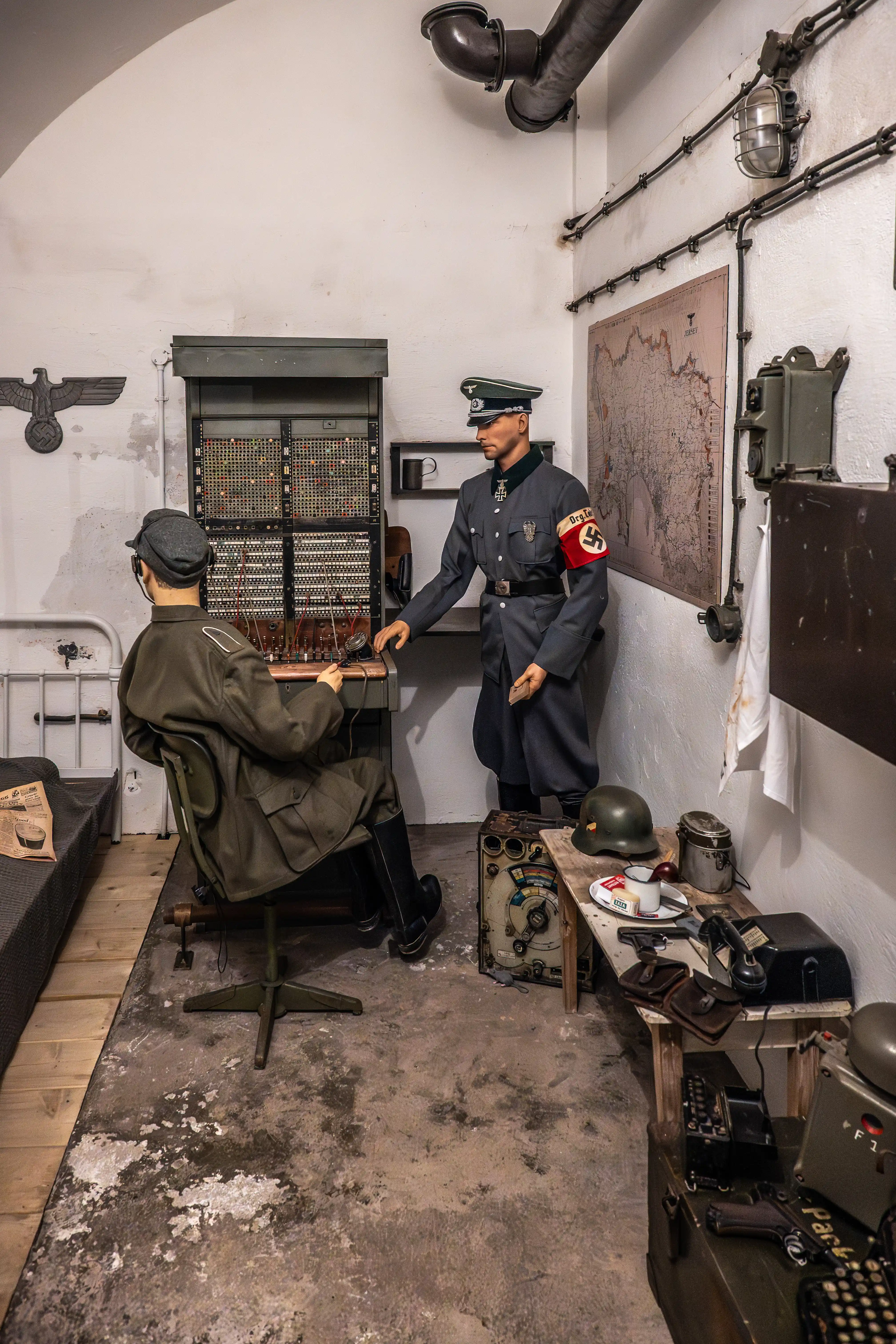 Two mannequins in World War II German uniforms are positioned in a replica command room. One mannequin, seated at a large switchboard, wears a dark green uniform and cap, while the other, standing, is in a grey uniform with an armband, looking towards the switchboard. The room is equipped with a map, telephones, helmets, and other period items.