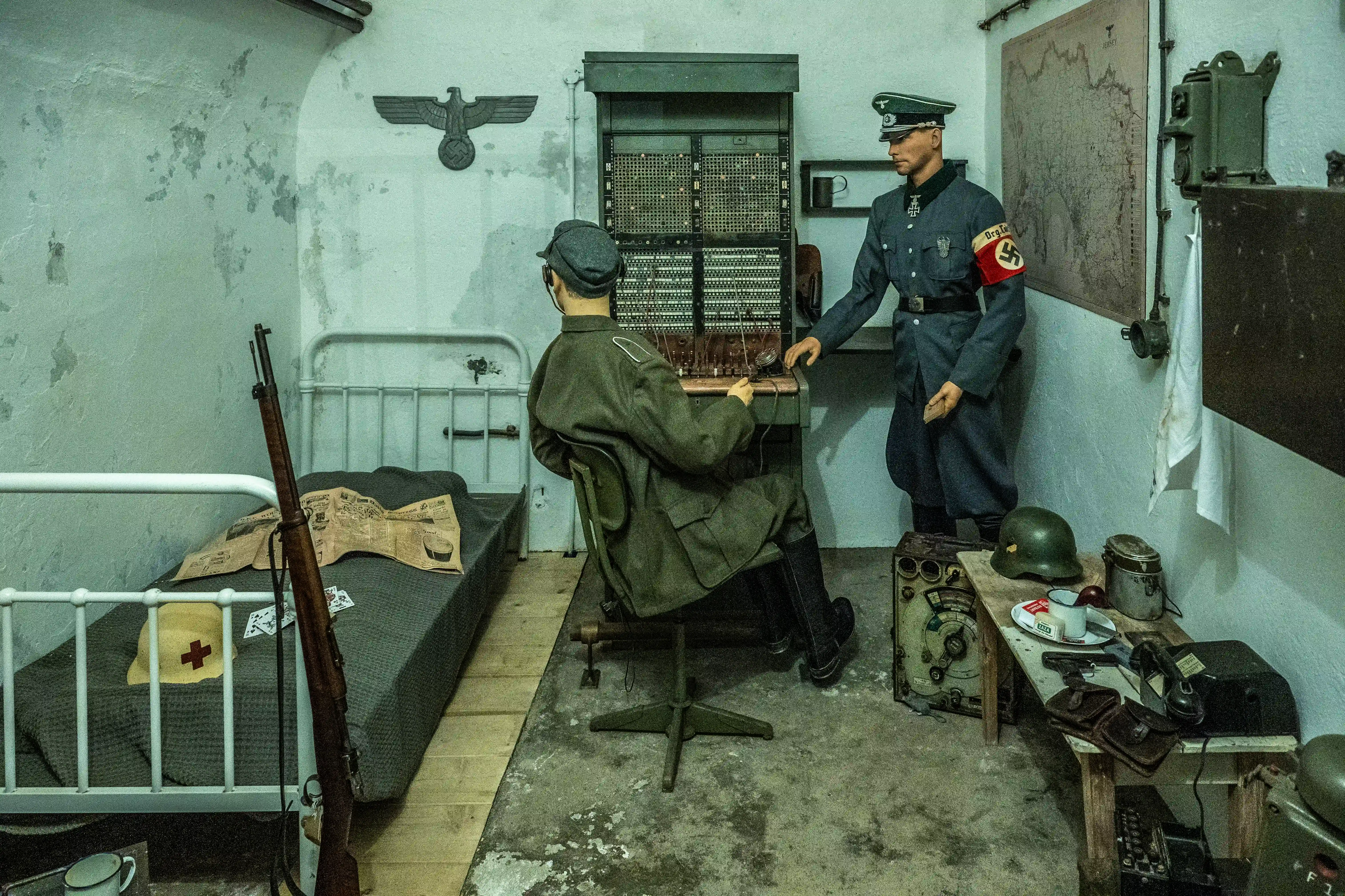 A scene inside a German communications bunker during World War II, featuring mannequins in uniform operating a switchboard, a military bed with a rifle, and strategic maps on the wall.
