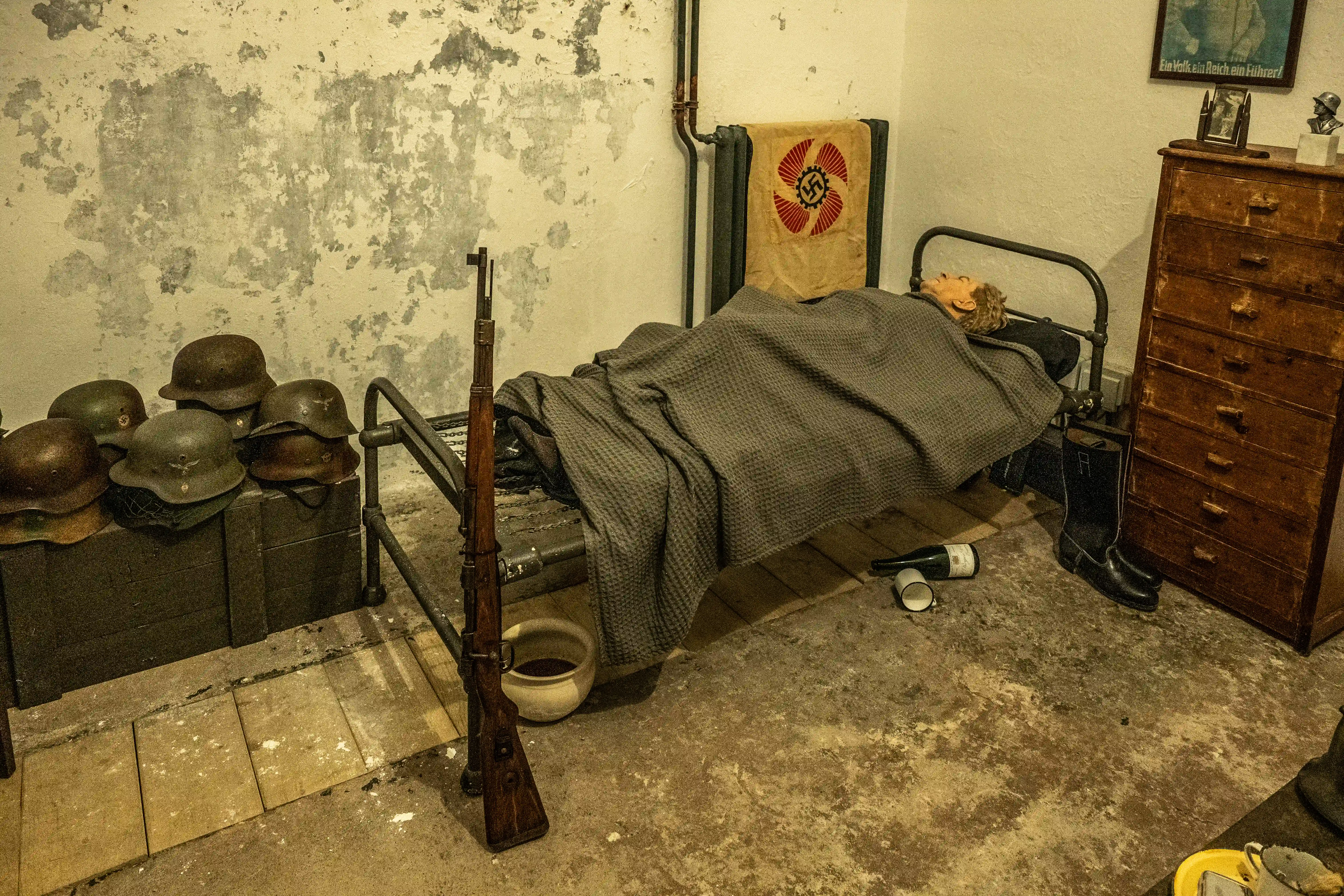A dimly lit, staged room showing a mannequin in a military-style bed, surrounded by German WWII helmets, a rifle, a Nazi flag, and a wooden dresser with boots.