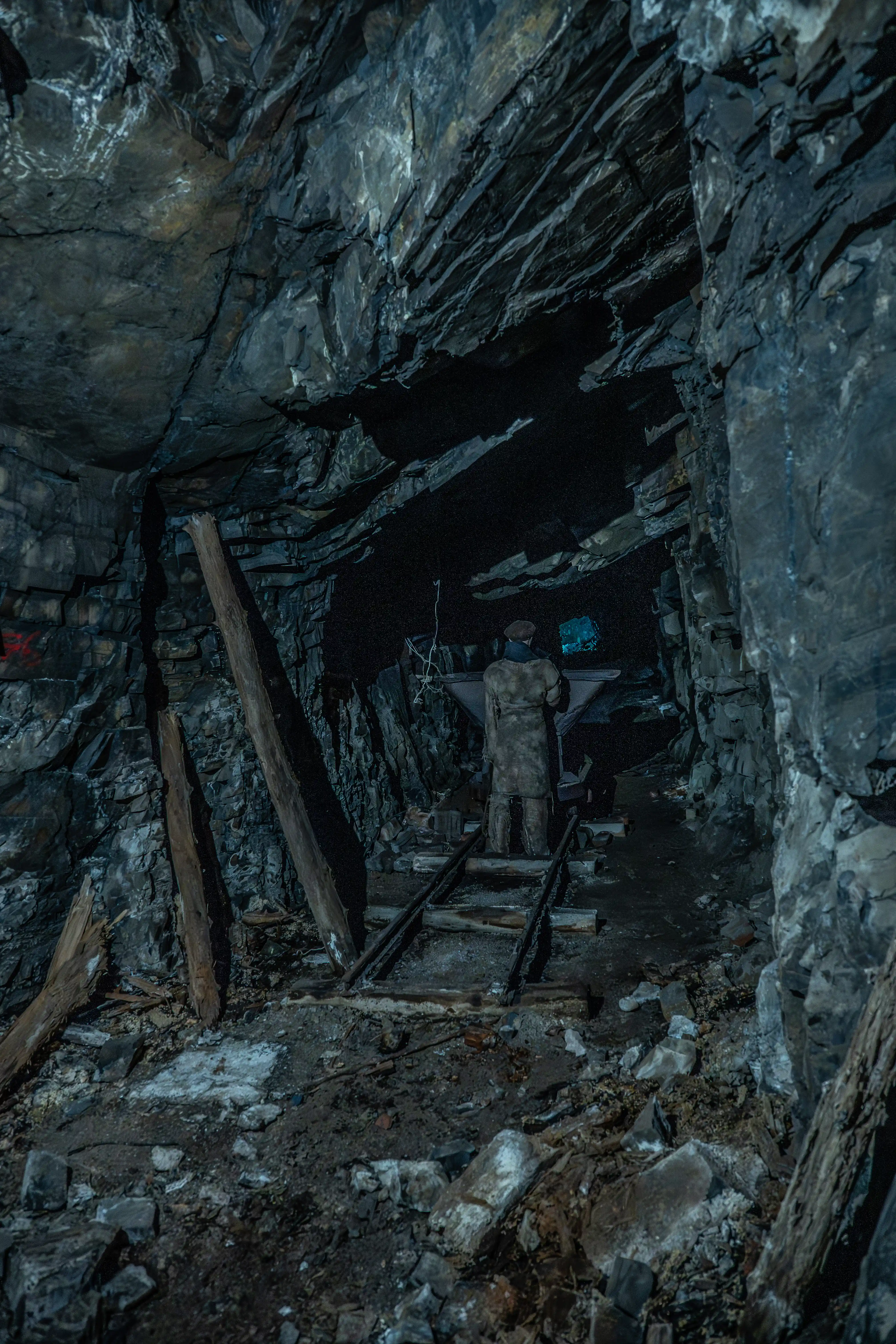  A person in a miner's outfit stands next to a mine cart on railway tracks inside a dark, rocky mine tunnel, with a blue light visible in the distance.