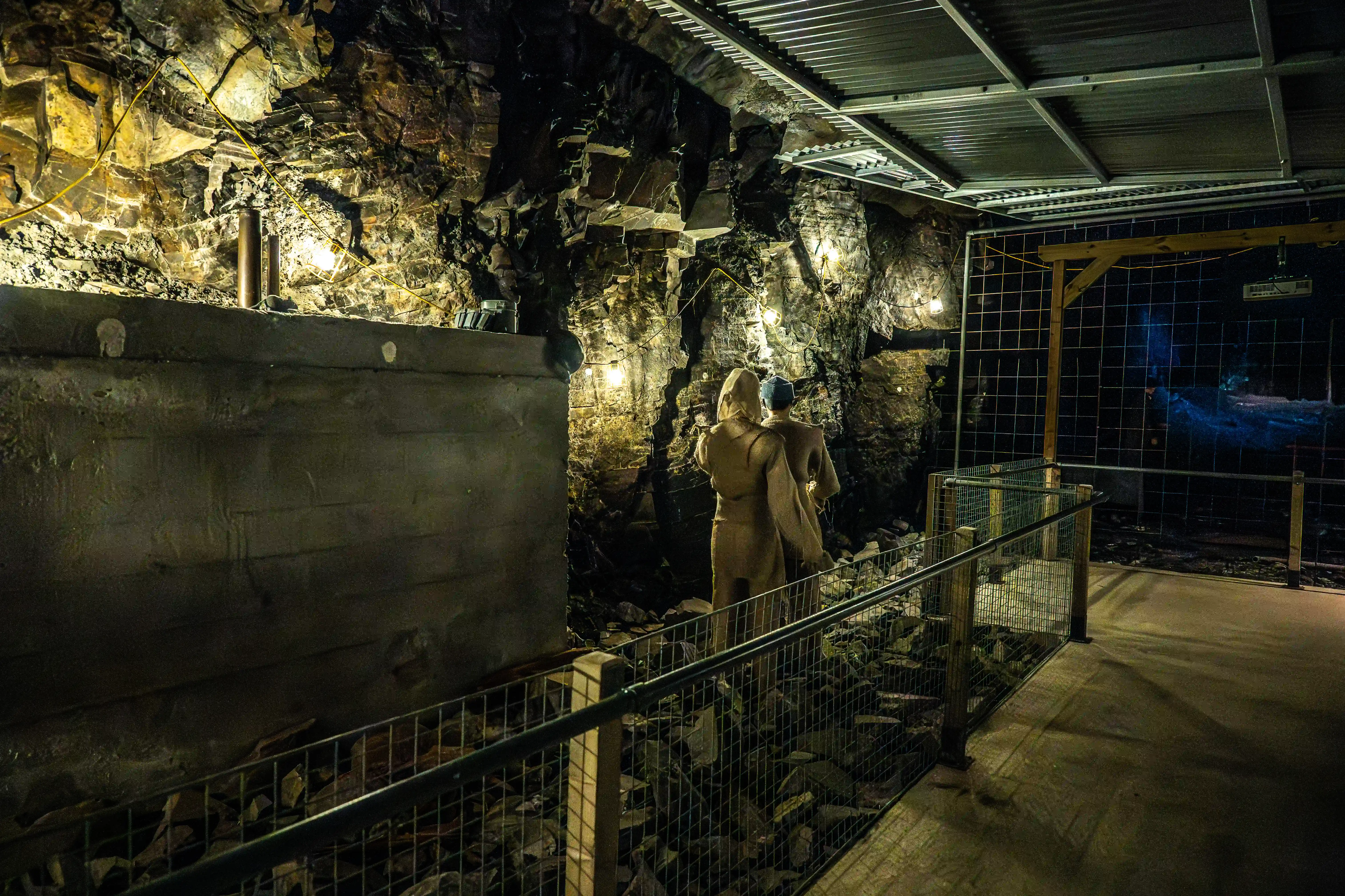 A dimly lit cavern with rough rock walls, featuring two mannequins in the foreground. Metal fencing and a walkway are visible, with lights casting shadows on the floor.