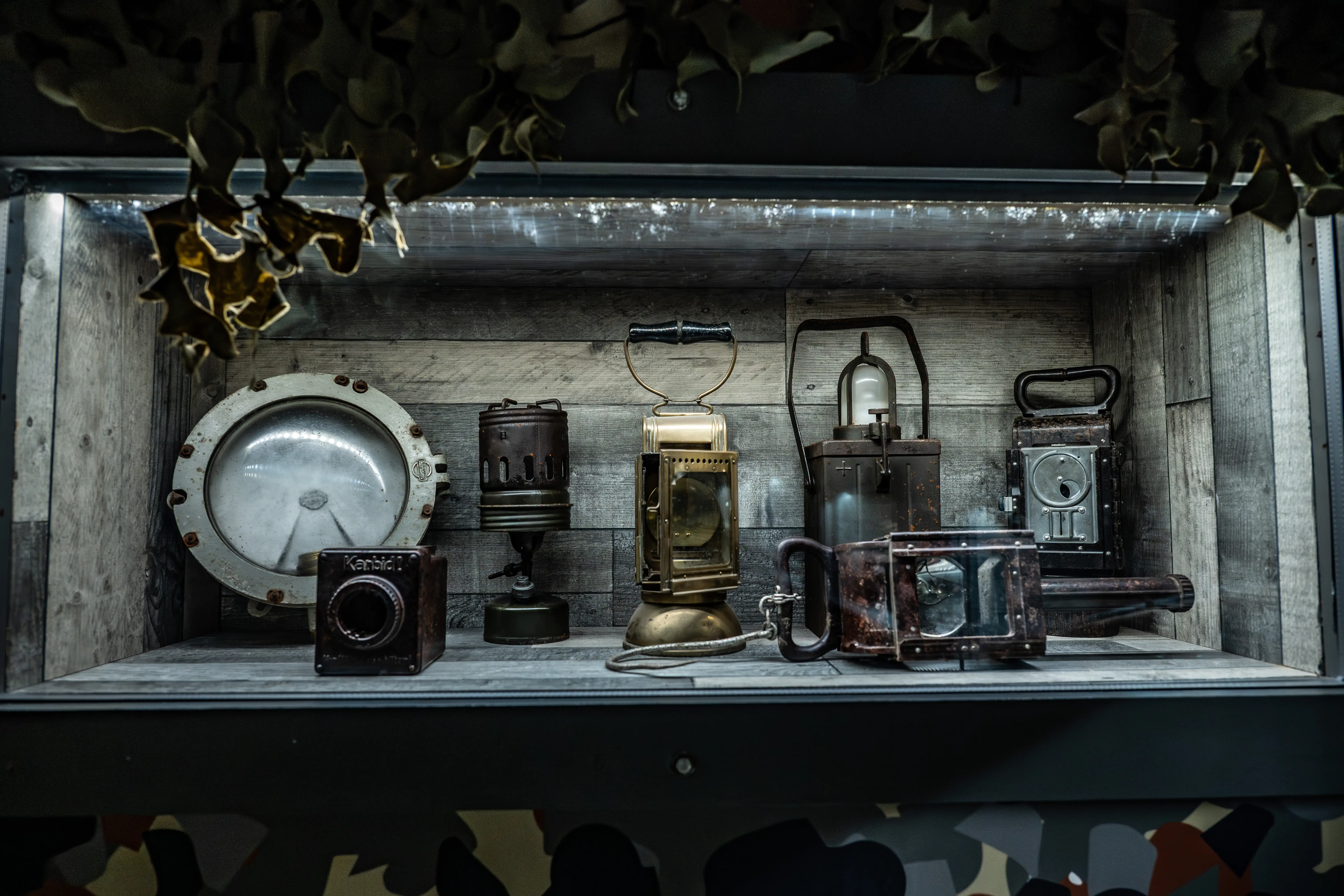 A dimly lit display shelf featuring a collection of antique lanterns, a large circular light, a small vintage camera, and other weathered metal equipment, with camouflage netting hanging above.