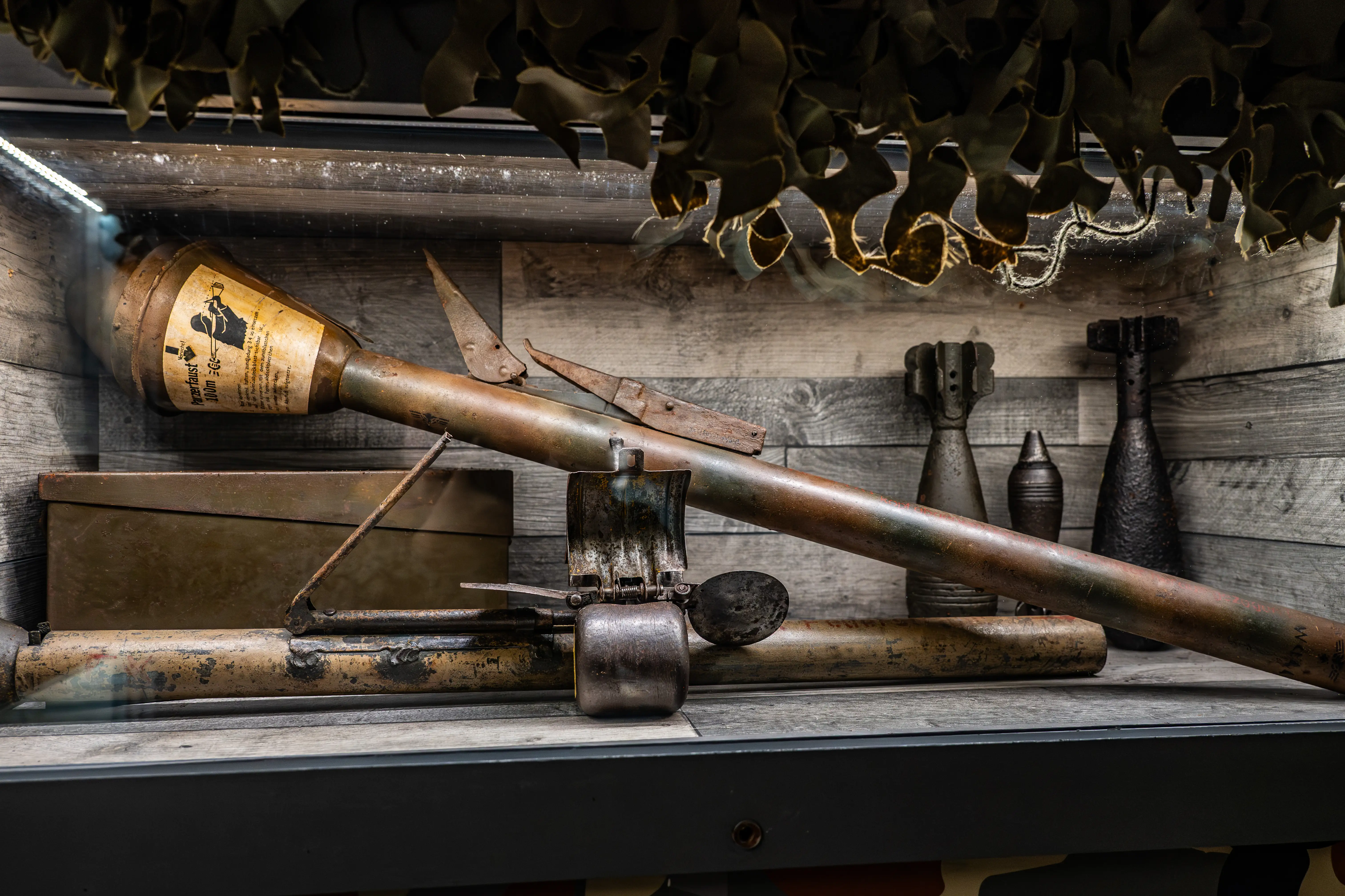 Close-up of a vintage Panzerfaust anti-tank weapon and several mortar shells arranged on a wooden shelf, with a camouflage net draped above