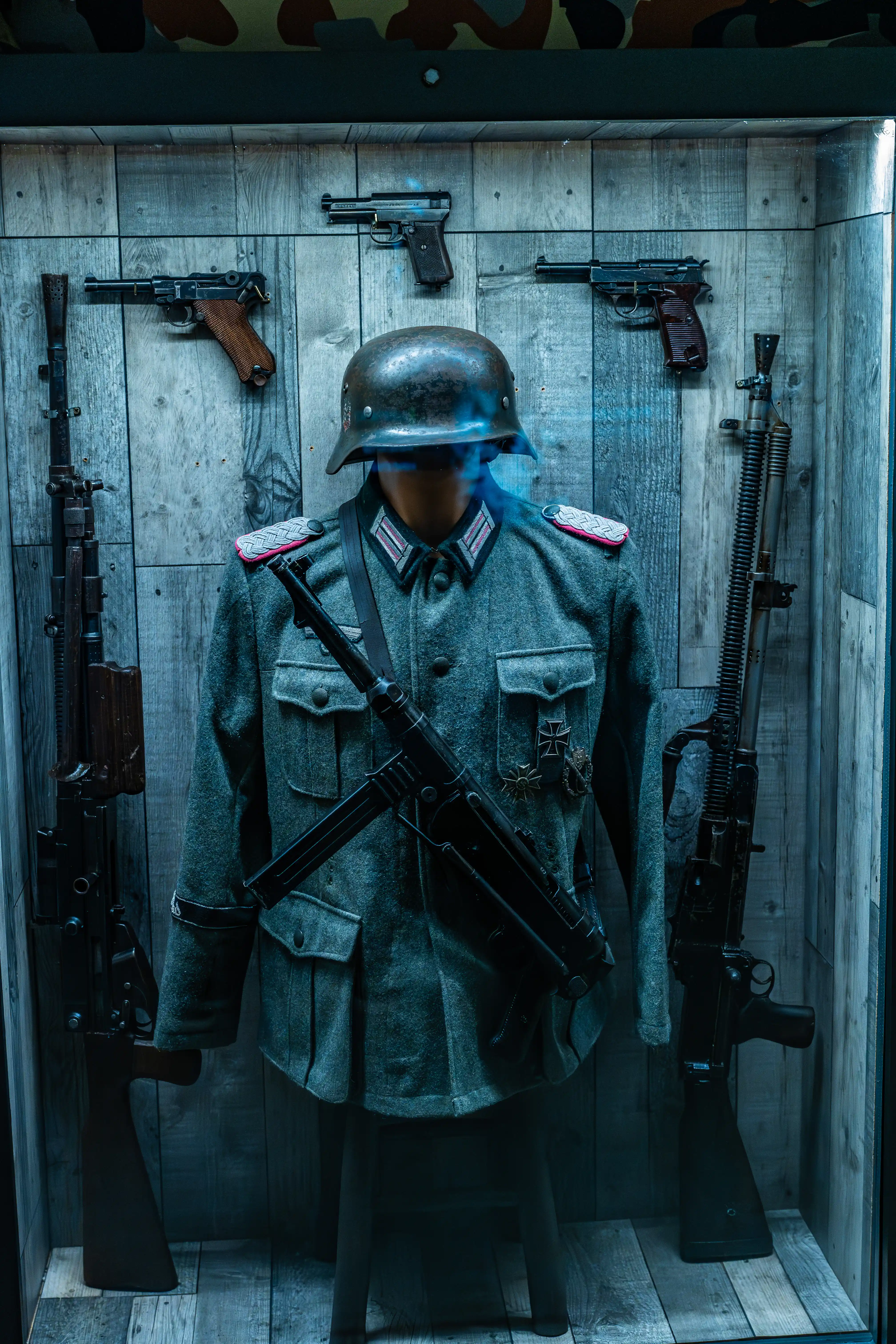 A mannequin dressed in a World War II German military uniform, including a steel helmet and tunic with medals, surrounded by various firearms.