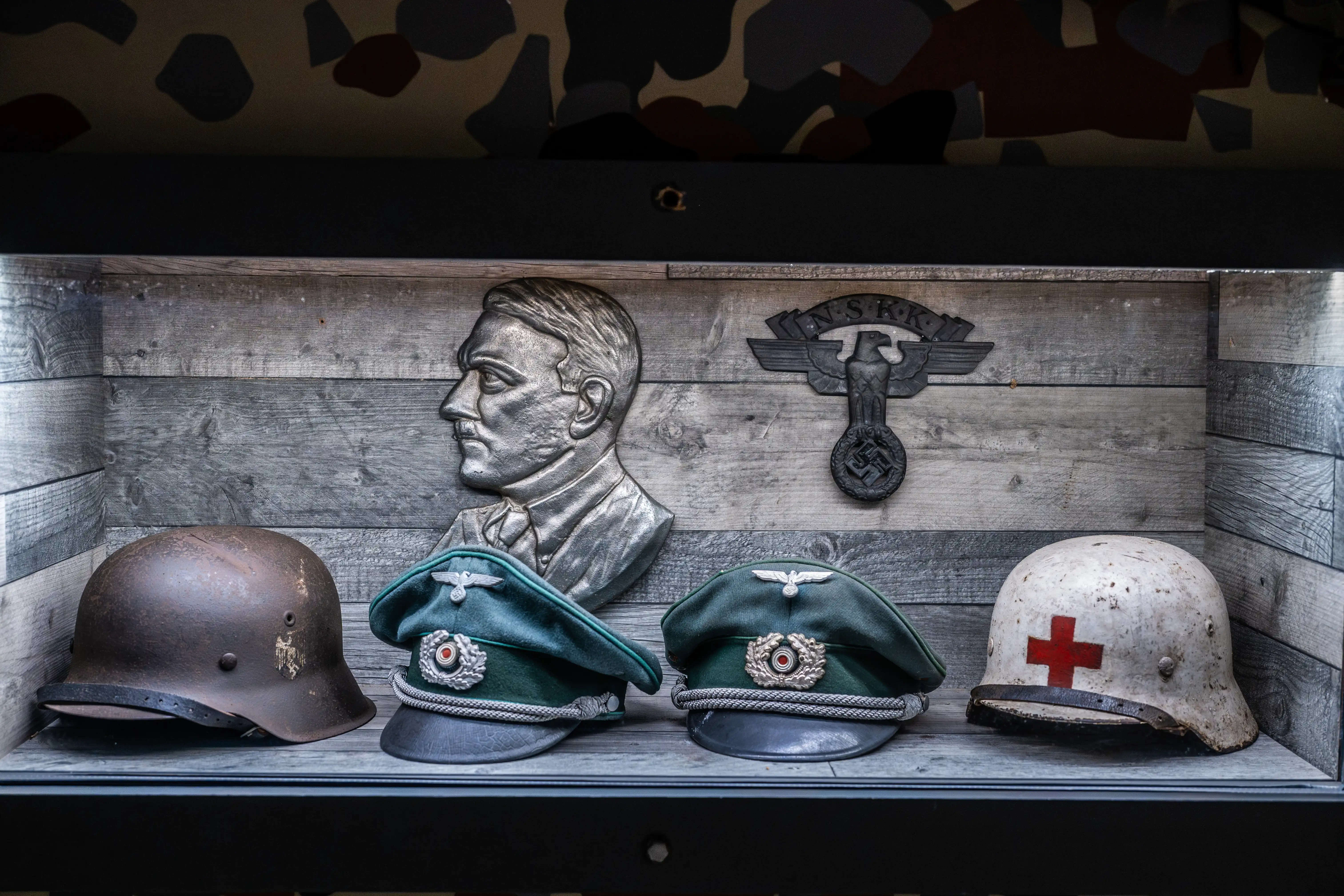 A display case containing a bust of Adolf Hitler, a Nazi emblem, two German military caps, and a white helmet with a red cross.