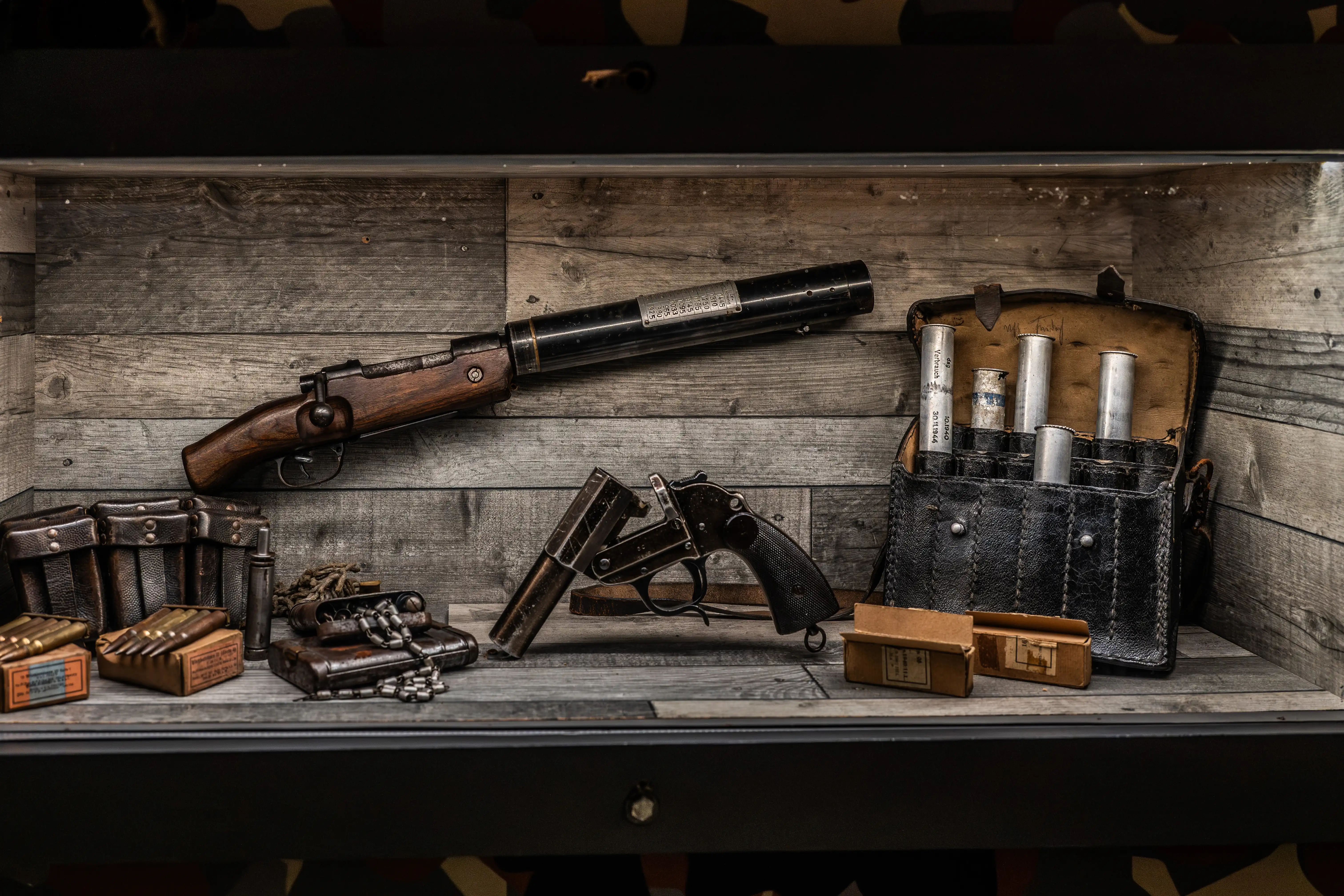 A wooden shelf displays a collection of vintage firearms, including a rifle with a long barrel attachment, a small break-action pistol, and several leather ammunition pouches filled with cartridges and other accoutrements. The background is made of weathered wood panels.