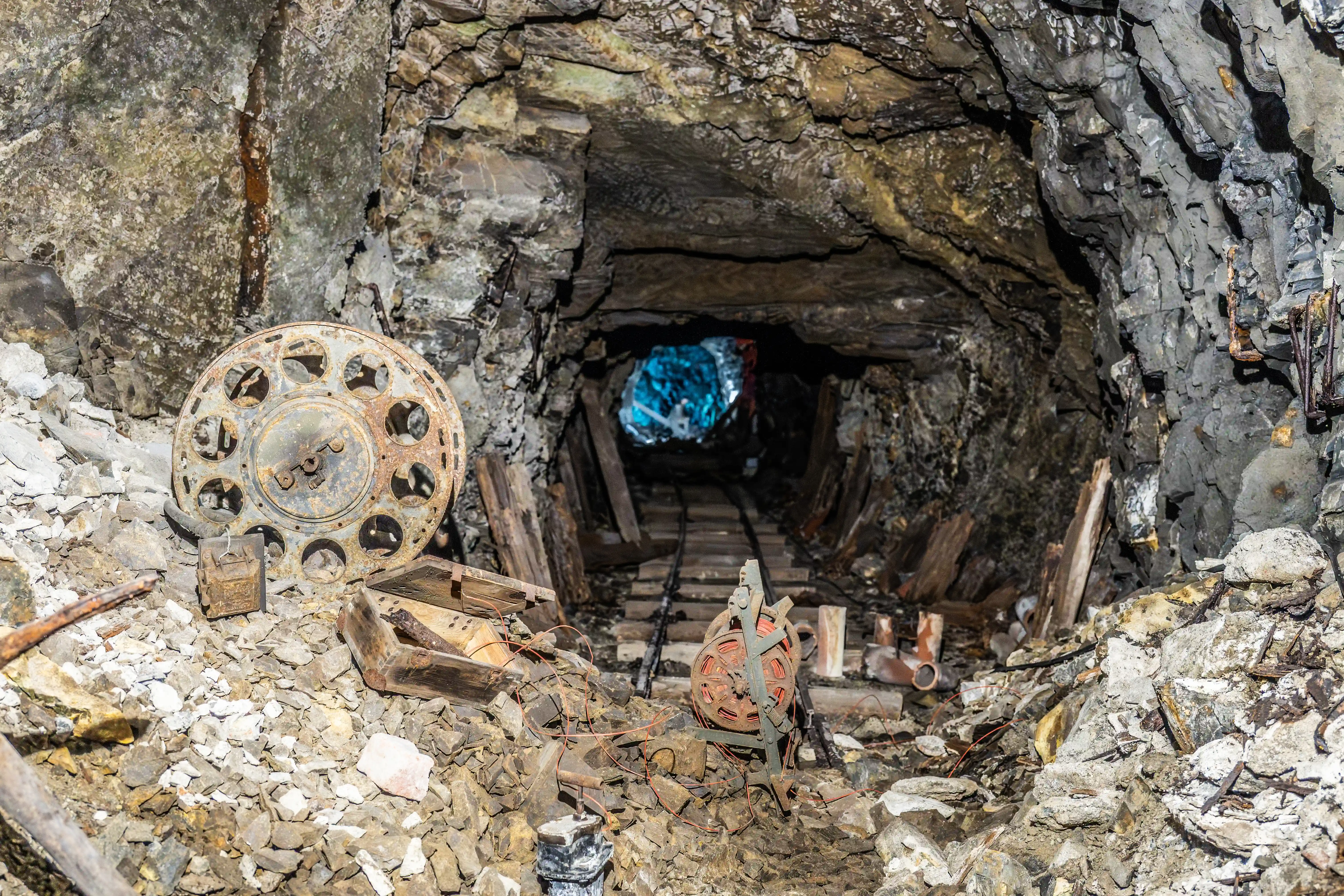A dark, abandoned mine tunnel with timber supports and a railway track leading into the distance. Rusty mining equipment, including reels and debris, are piled in the foreground..