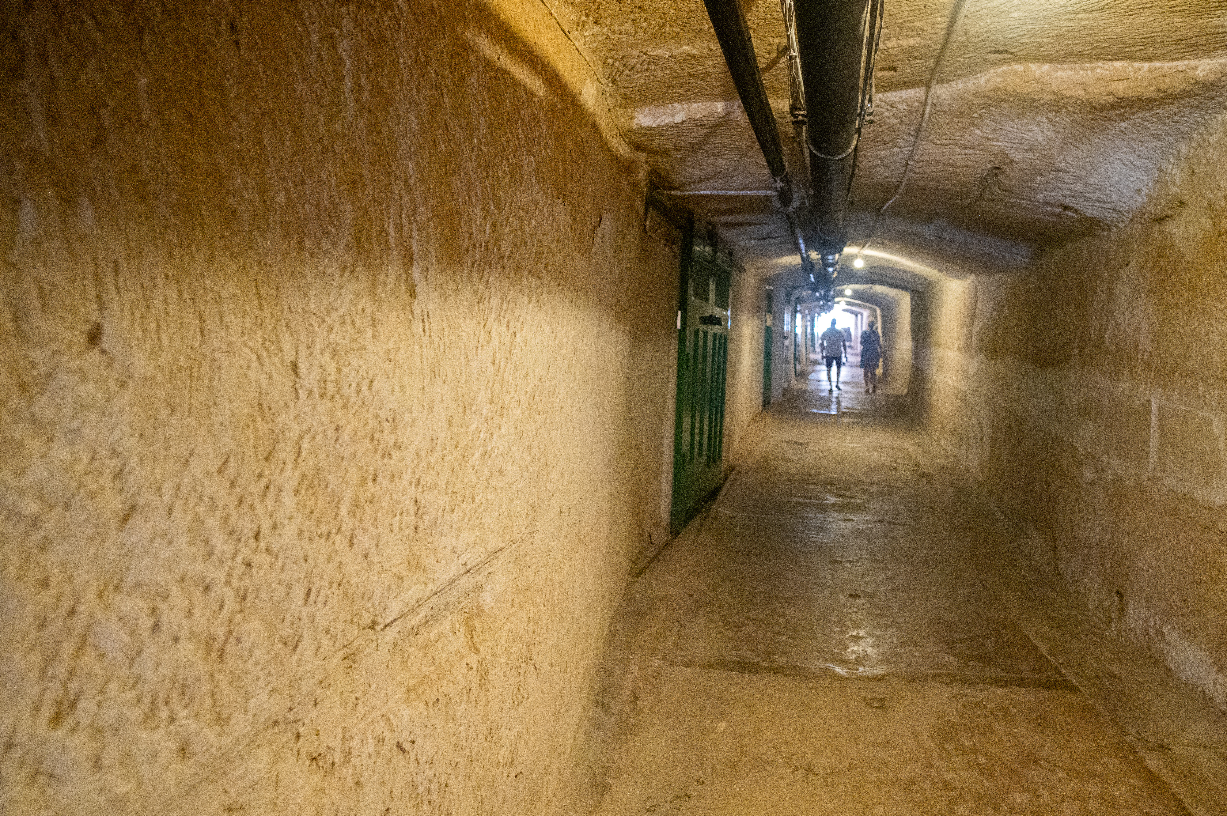 The tunnel is illuminated by overhead lights, and there are several green metal doors along the sides