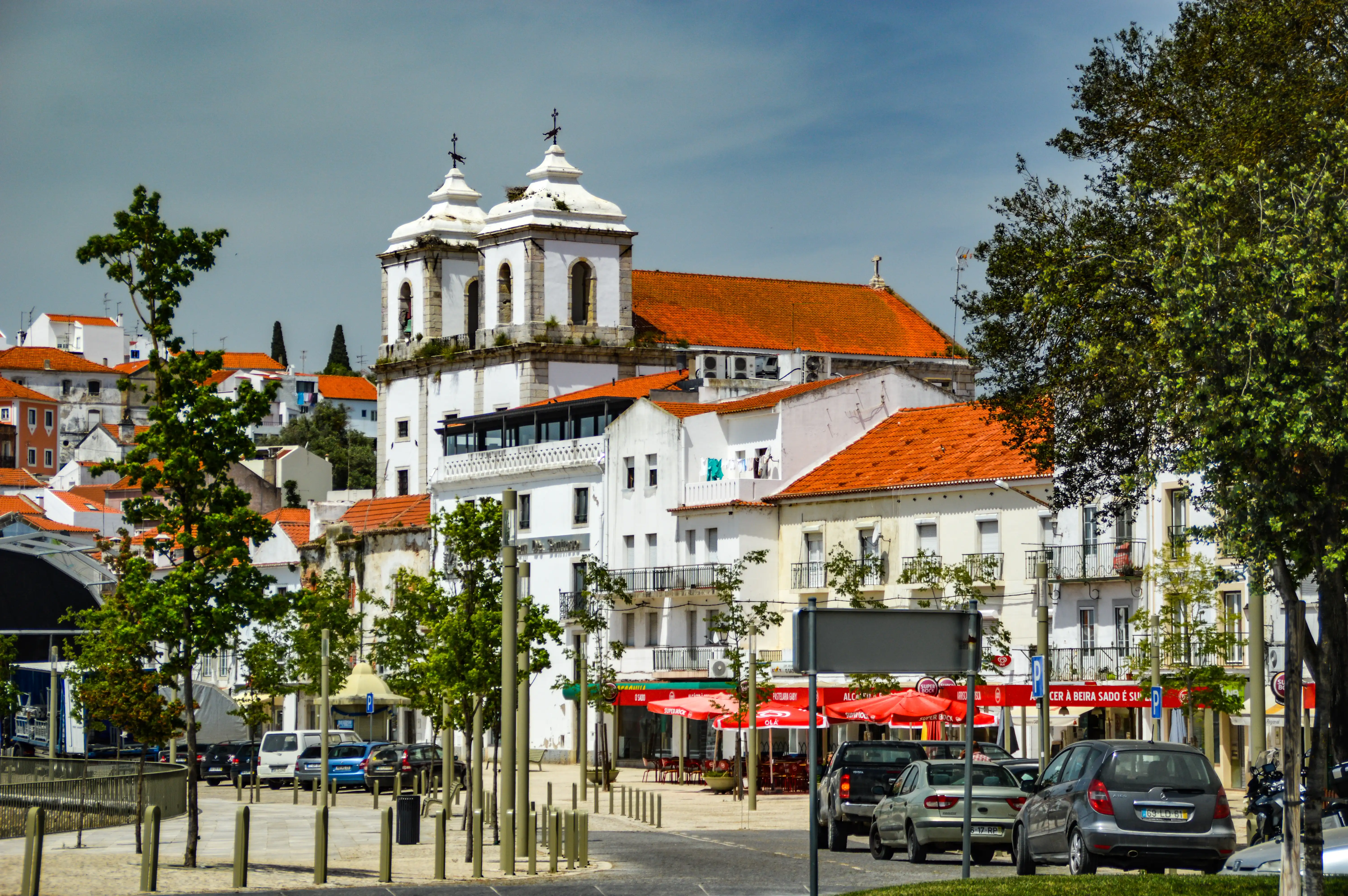 A picturesque view of a European town, showcasing a white church with two towers and red tiled roofs, with a street lined with cafes and parked cars in the foreground.