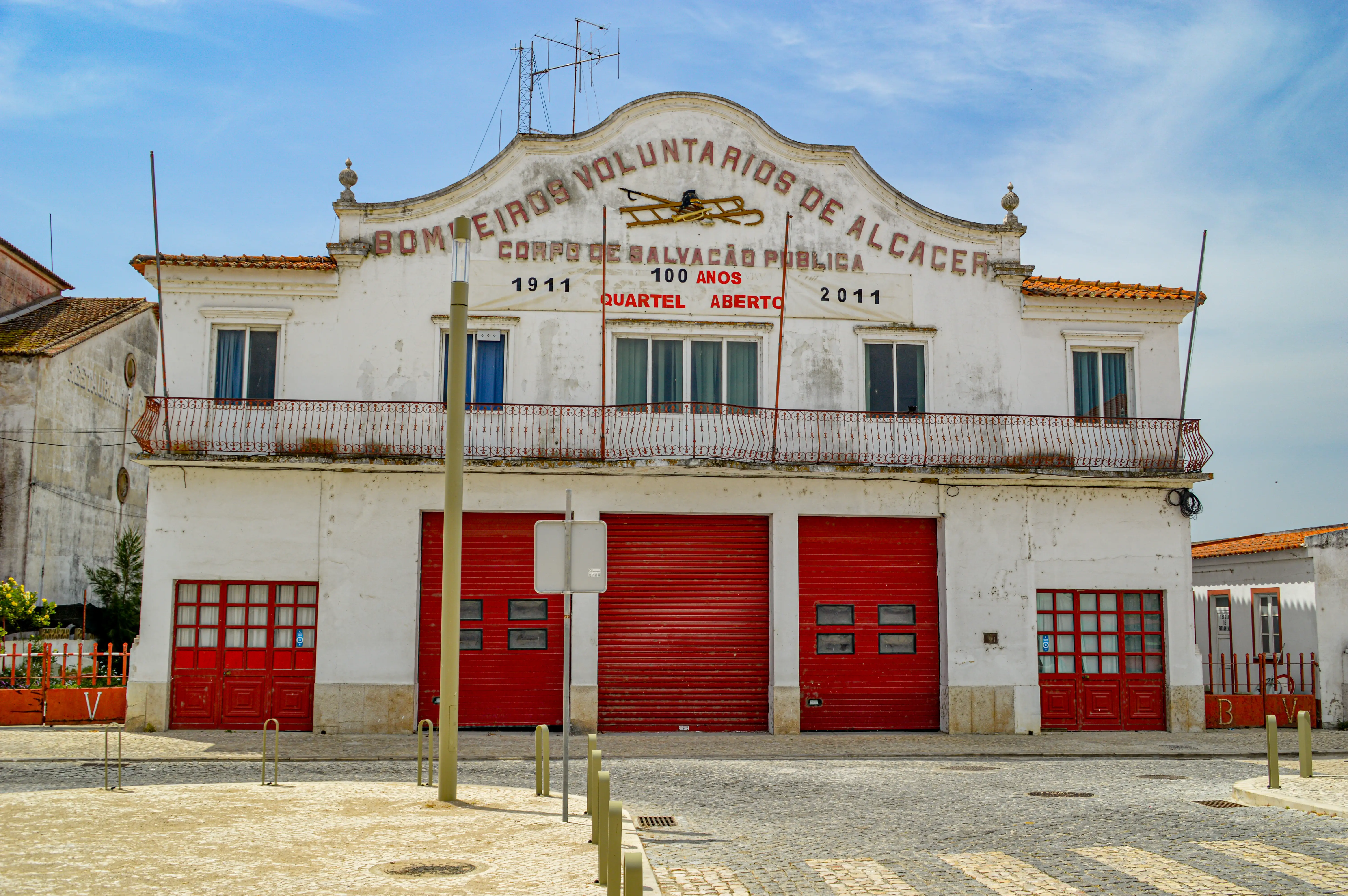 Exterior view of a white, two-story fire station building with red garage doors and a balcony, under a blue sky. Text on the facade reads Bombeiros Voluntarios de Alcacer and indicates a 100-year anniversary from 1911 to 2011.
