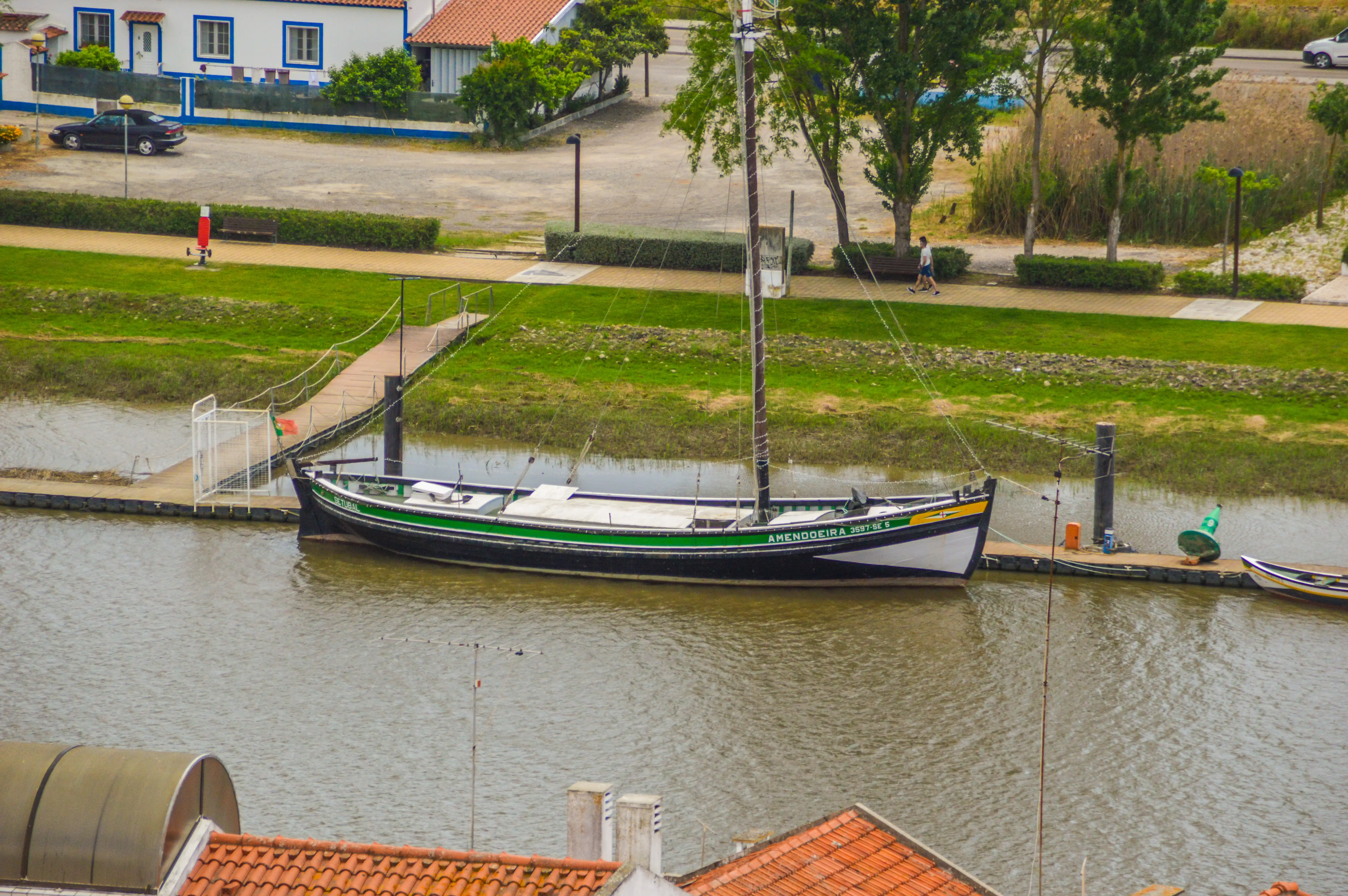 A black and green sailboat named AMENDOIEIRA is moored on a calm river next to a wooden pier. A paved path with green grass and bushes runs alongside the river. A white building with blue trim and a black car are visible in the background.
