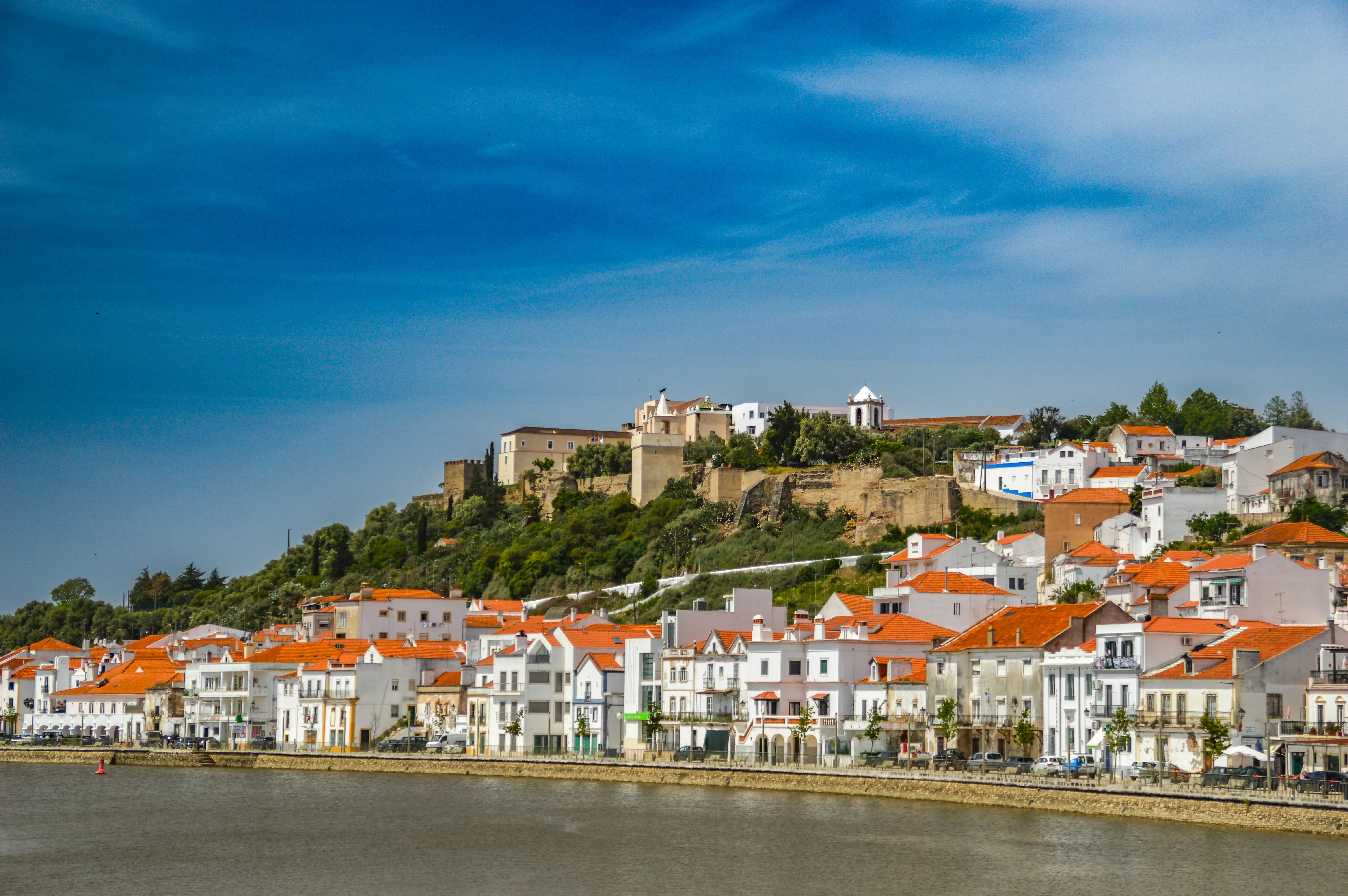 A waterfront view of a historic town with whitewashed buildings and terracotta roofs, overlooking a river, with a castle and church on a hill in the background under a blue sky.