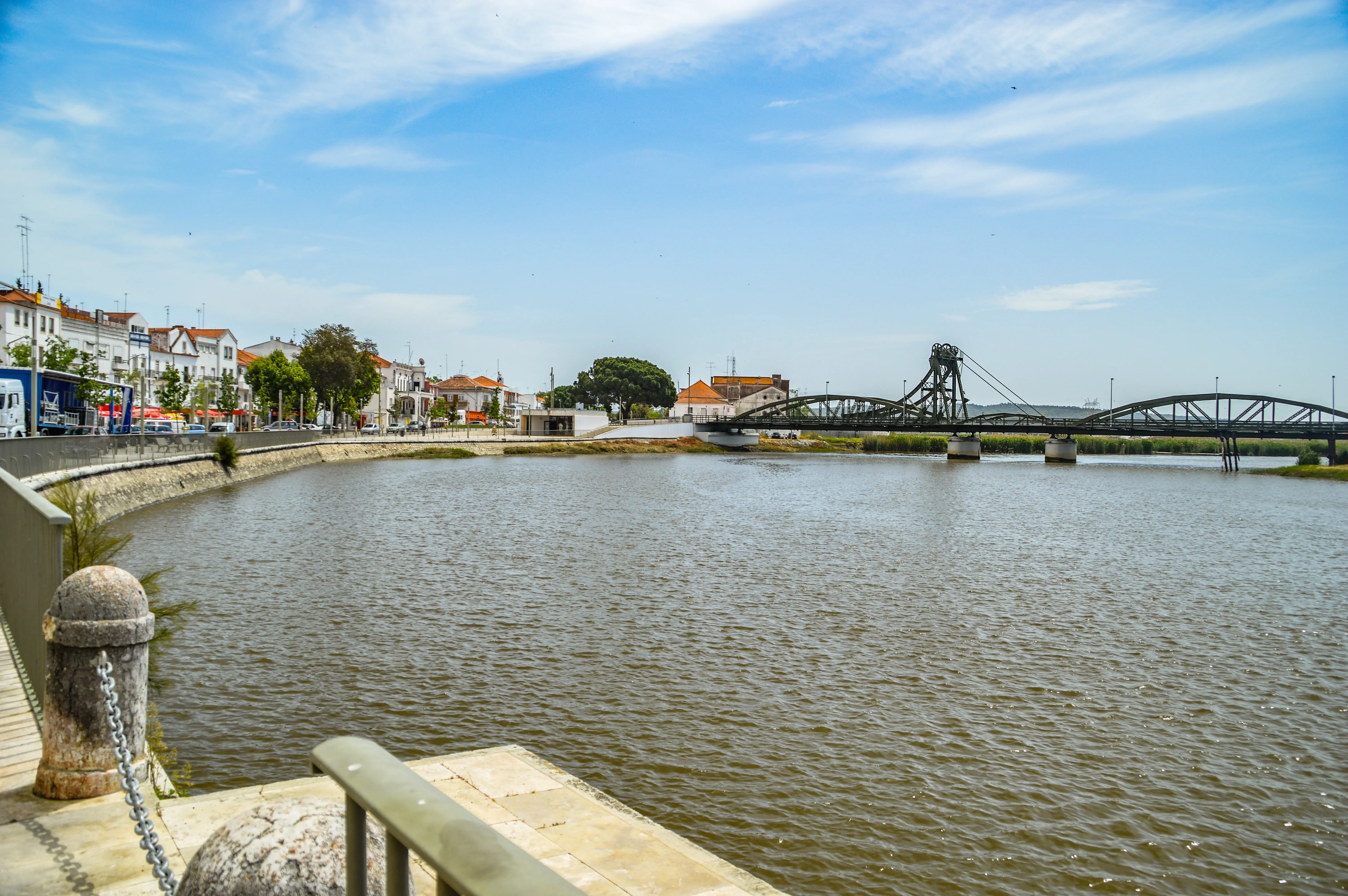  A wide river with rippling water is in the foreground. Along the left bank are buildings with white walls and orange tile roofs. A green metal bridge with a tall central structure crosses the river in the distance. The sky is blue with some white clouds