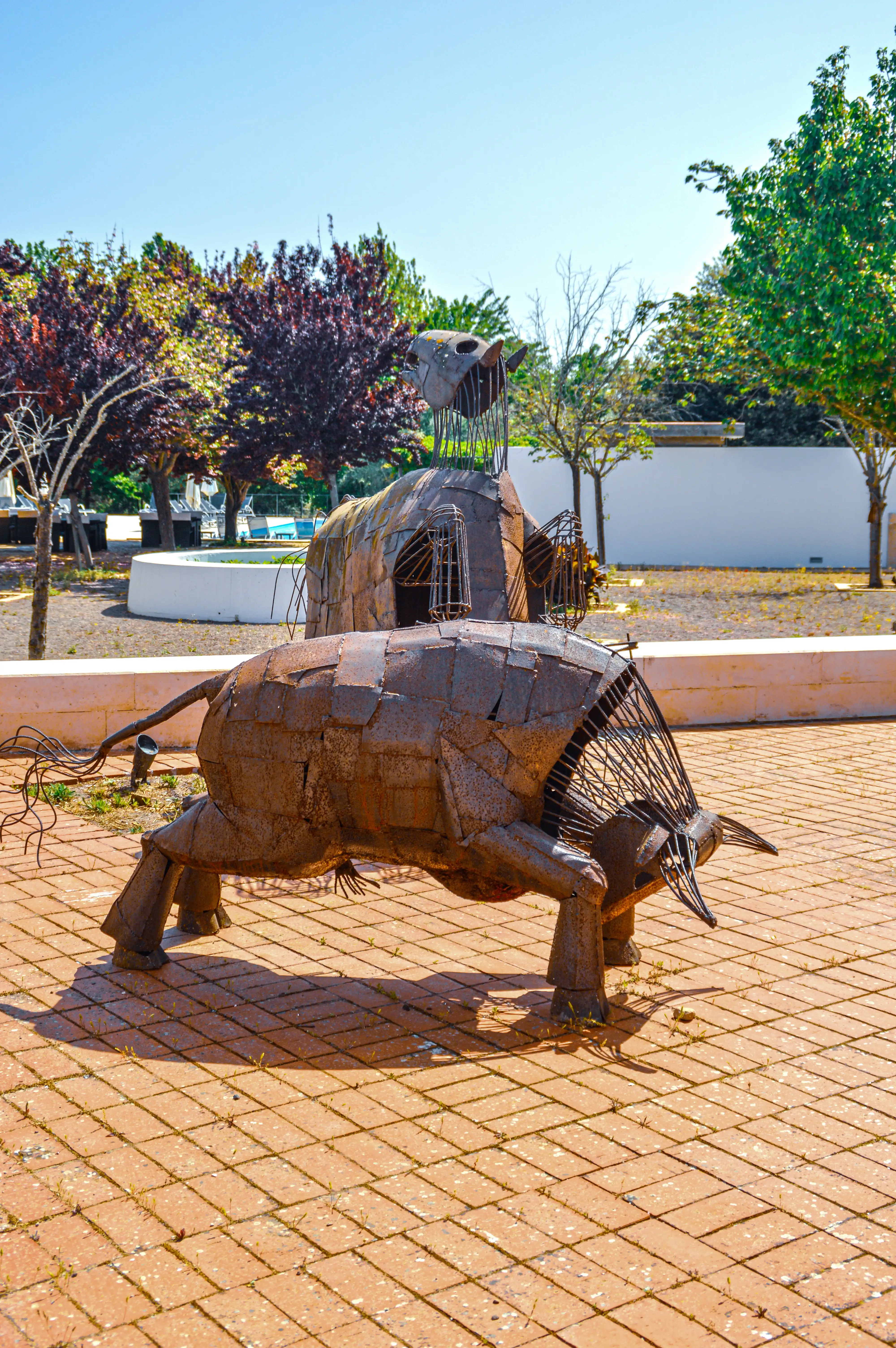 A metallic sculpture of a bull, made from rusty metal plates, is positioned on an orange brick pathway. The sculpture is in the foreground, with trees and a white wall in the background under a bright blue sky.