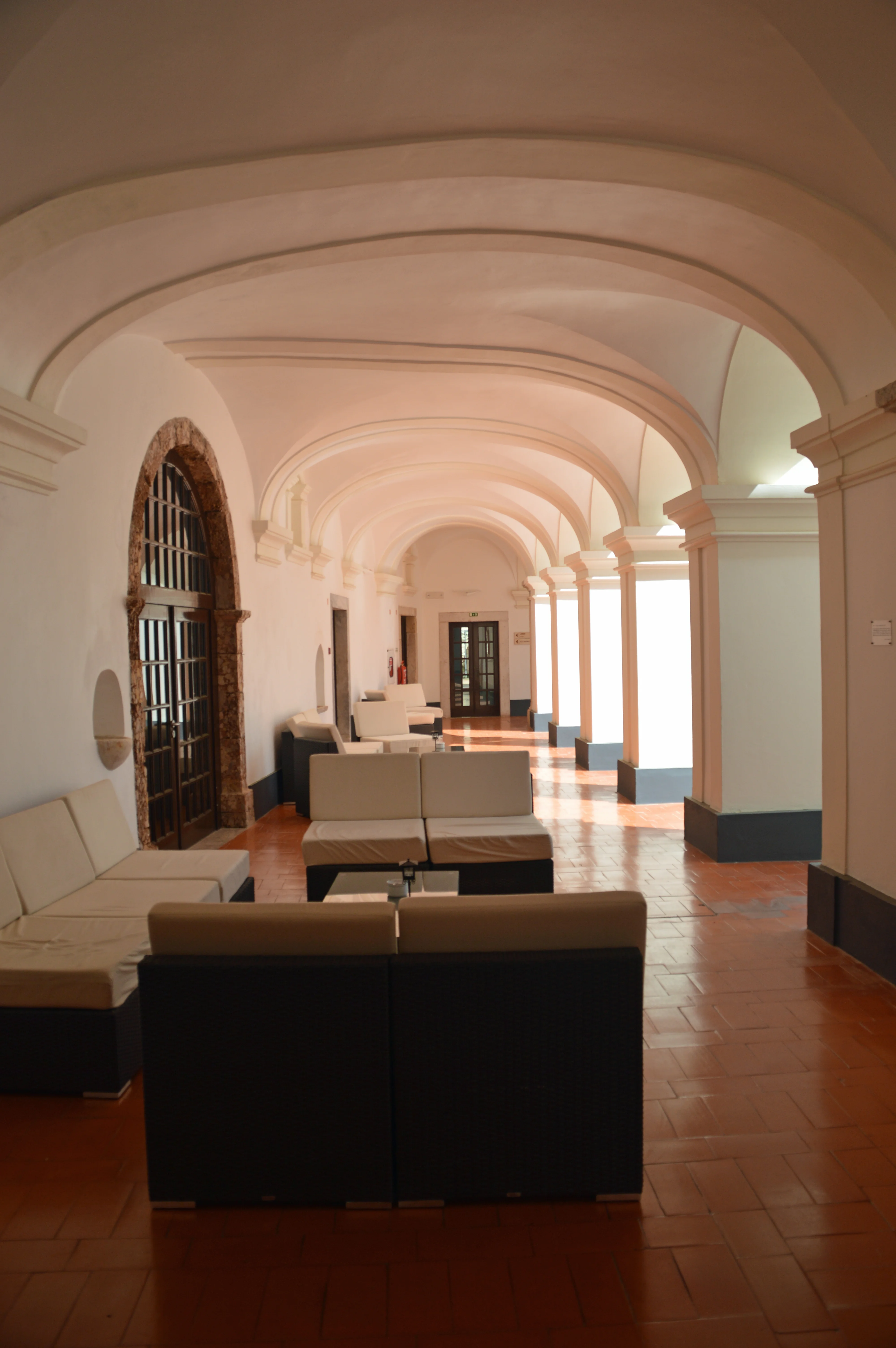 A spacious, arched corridor with white walls and a terracotta tiled floor. Contemporary cream sofas are arranged along one side, with darker modular seating in the foreground. The corridor recedes into the distance, illuminated by soft, warm lighting.