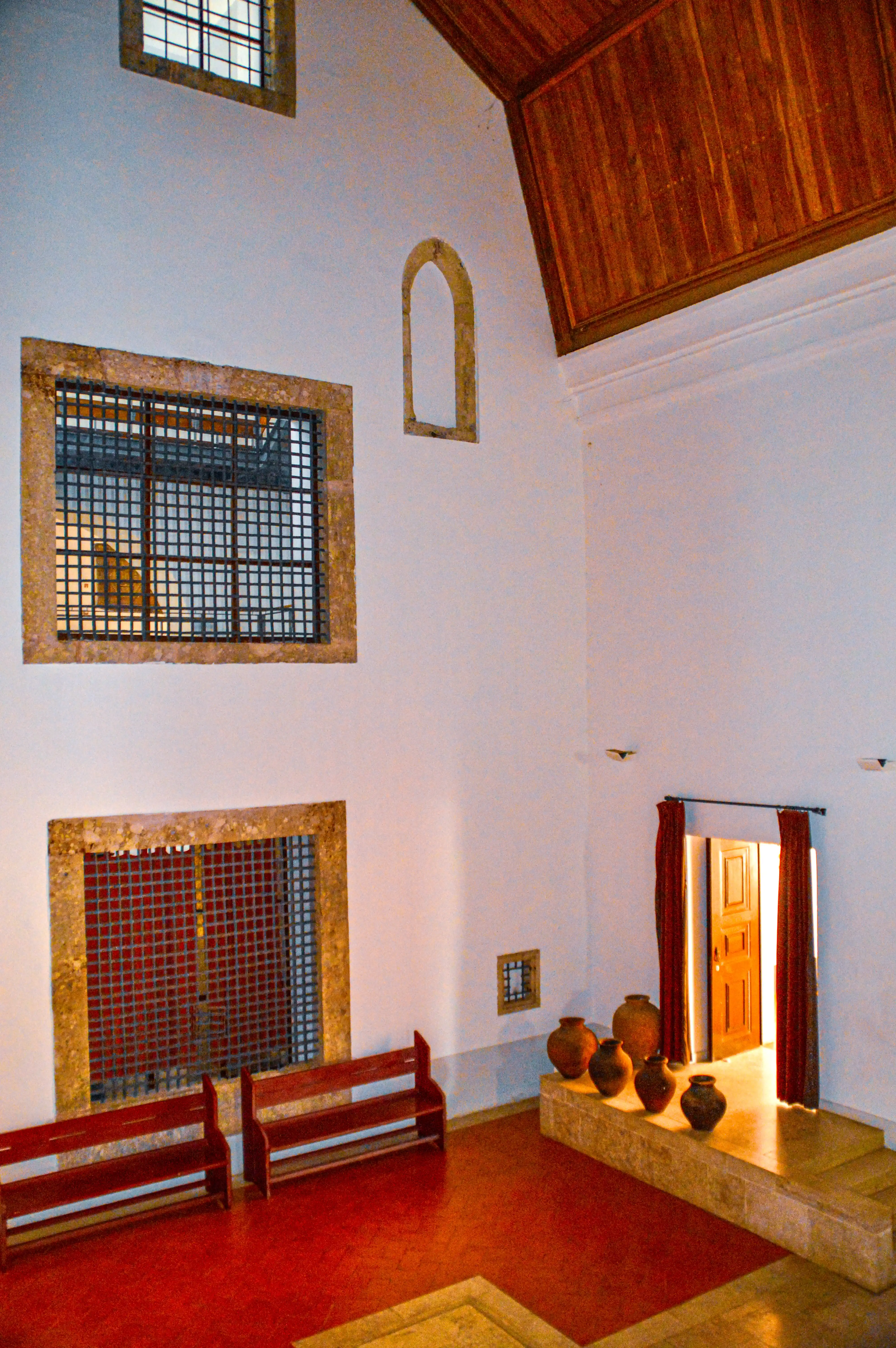 Interior of a room with white walls, a wooden ceiling, two windows with barred fronts, a red floor, two wooden benches, a raised platform with pots, and a doorway with red curtains.
