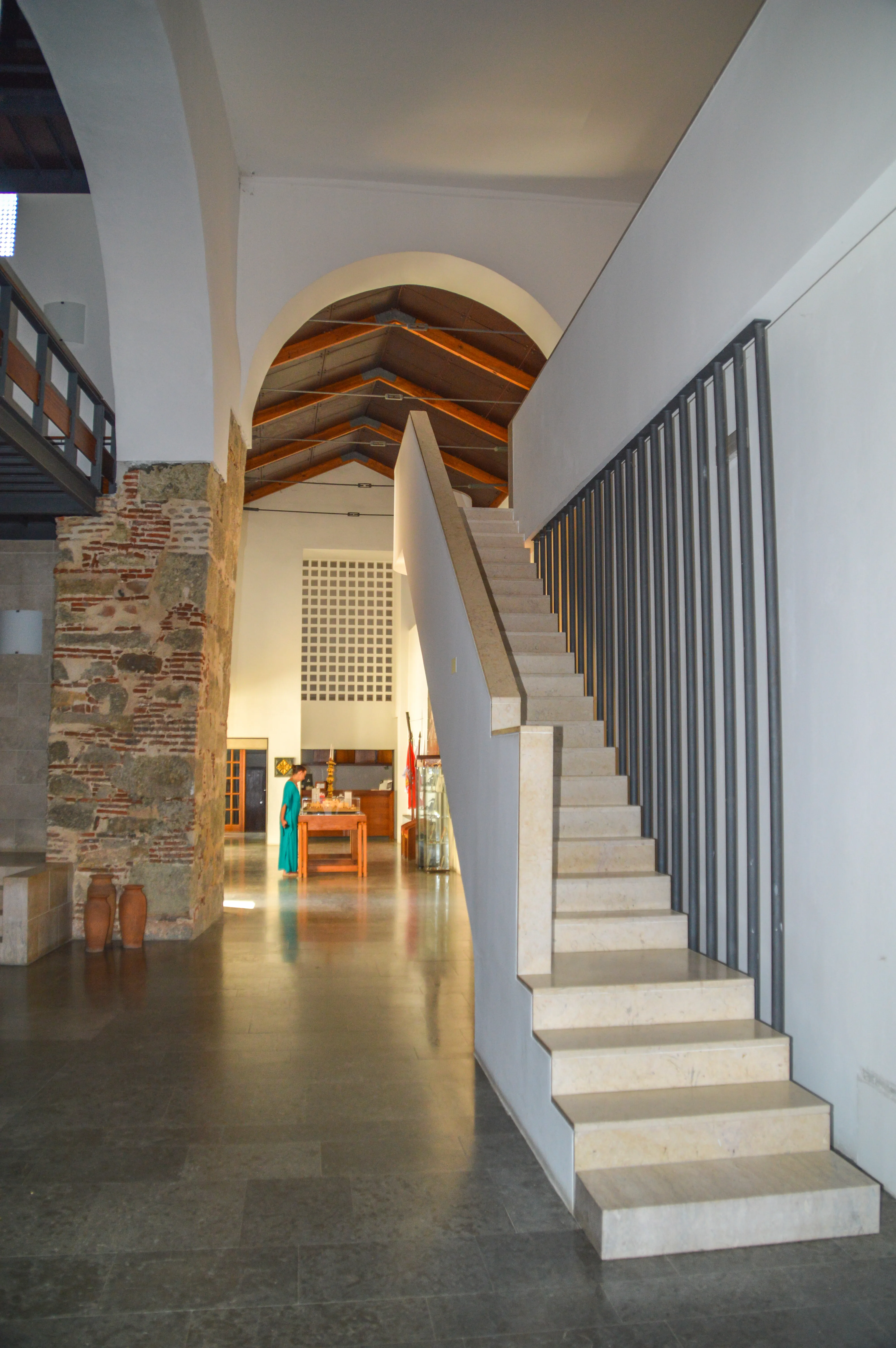 Interior of a building with a prominent white staircase and a dark metal railing. Exposed brickwork and stone are on the left, leading to an arched ceiling with visible wooden beams. In the distance, a person in blue stands by a wooden table.