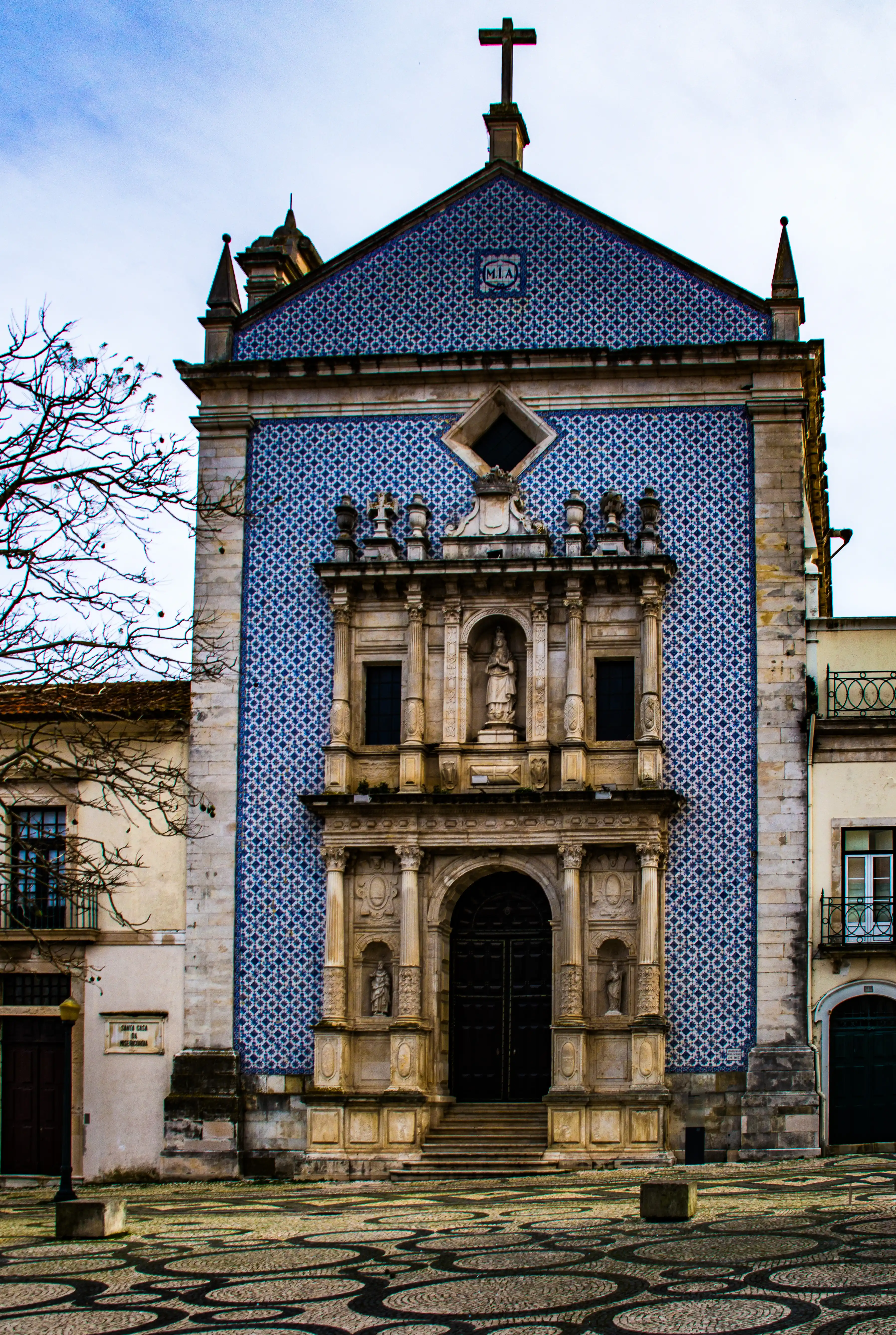 A church building with a large facade covered in blue and white geometric azulejo tiles, a stone carved entrance with a statue, and a cross on top. The ground is paved with decorative black and white cobblestones, and a person stands on the right.