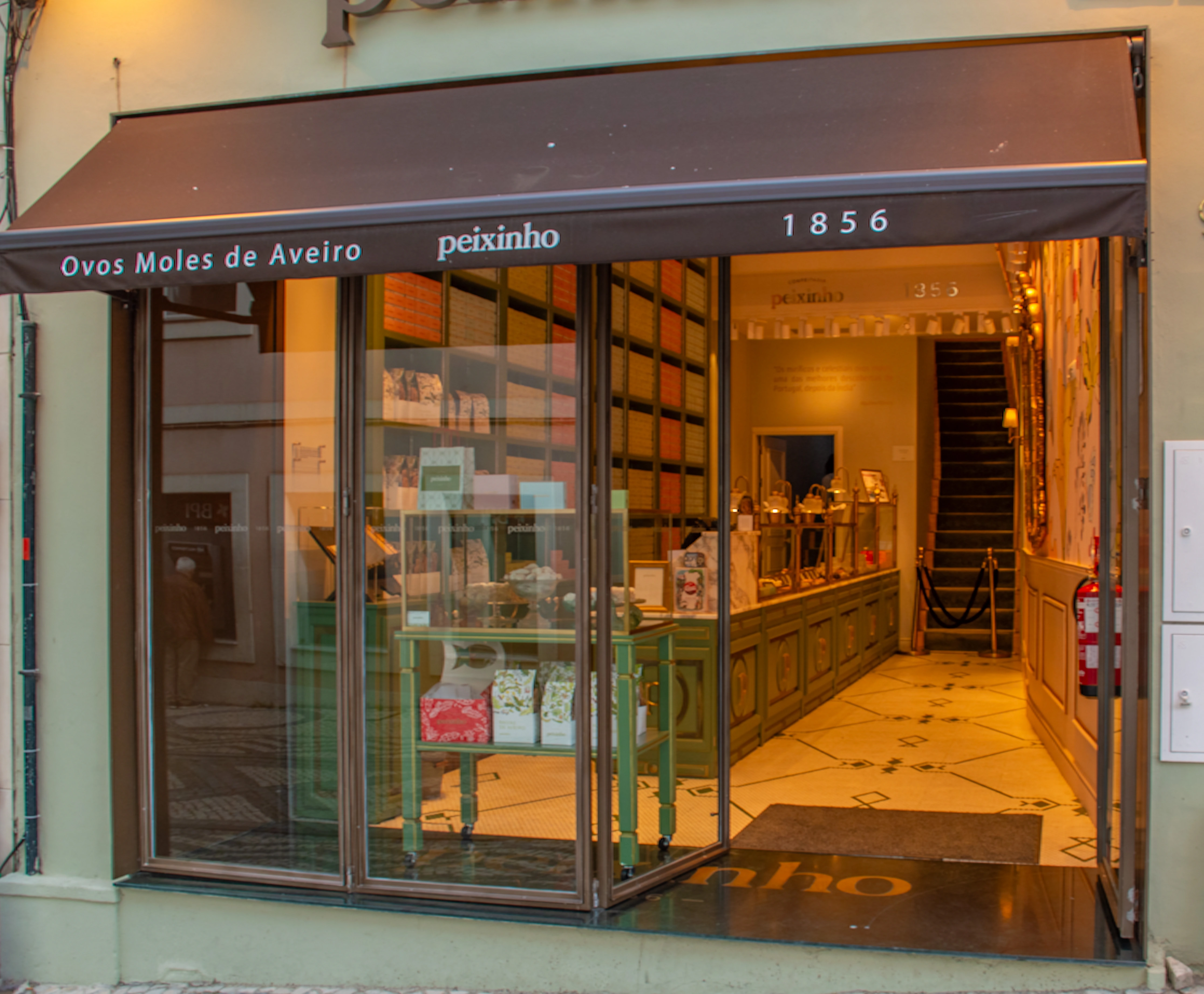 A brightly lit confectionery shop called Peixinho in Aveiro, Portugal, specializing in Ovos Moles. The shop window displays rows of colourful sweet boxes and decorative items, with the year 