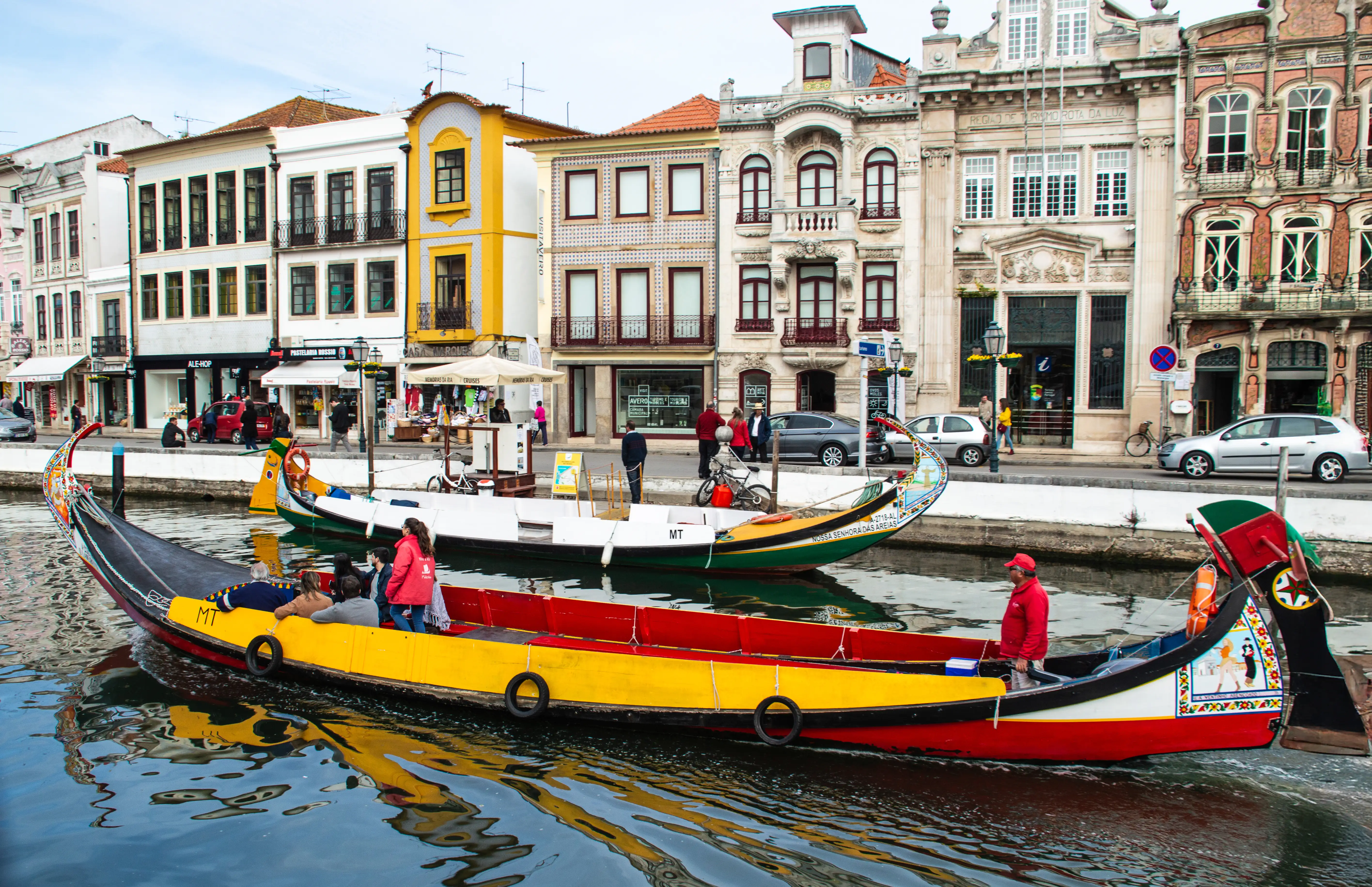 Two brightly decorated moliceiro boats, one predominantly red and yellow with passengers, the other green and white, float on a canal in Aveiro. Behind them, a row of ornate, pastel-coloured buildings line the waterfront.