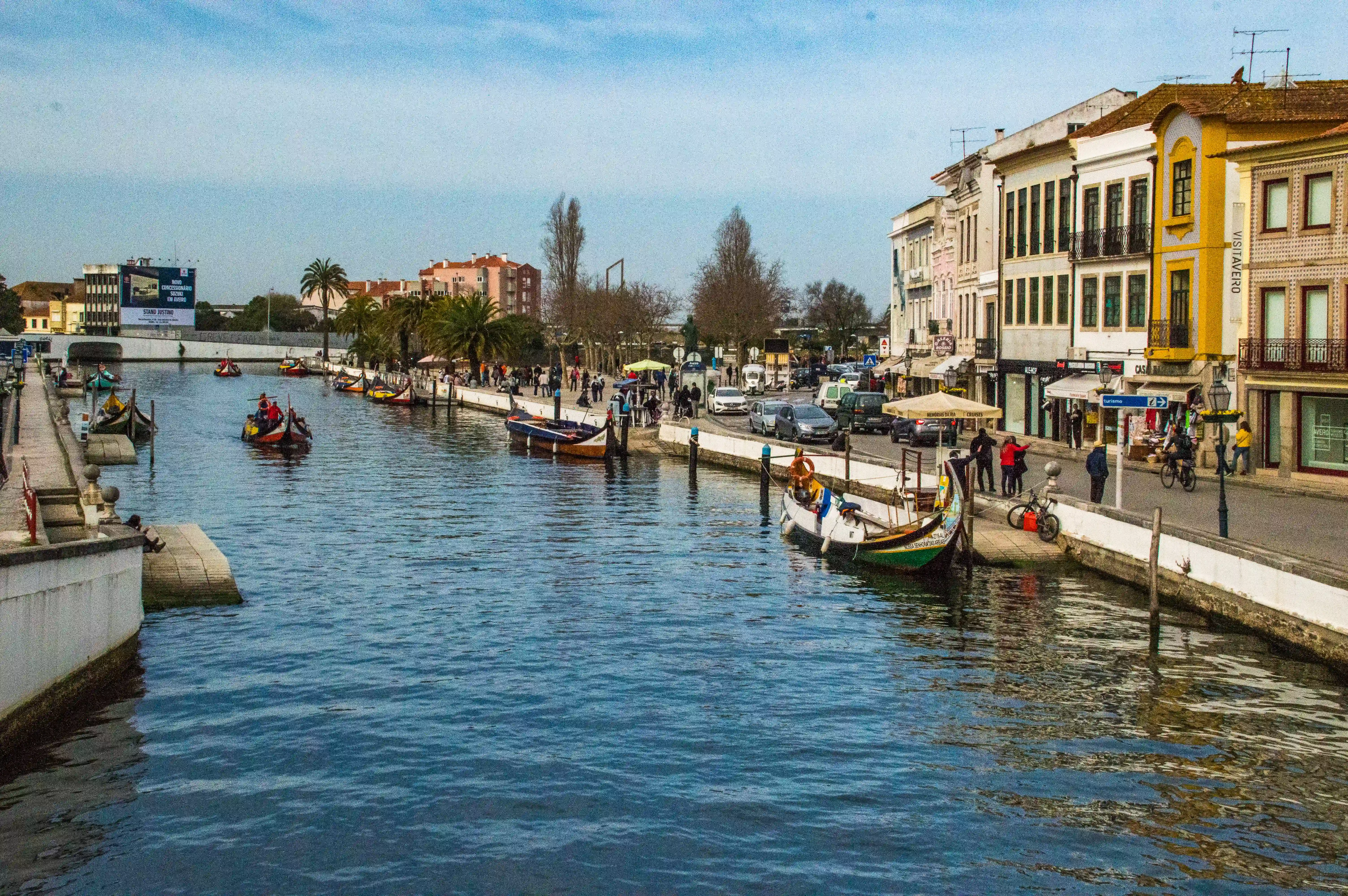 A tranquil canal scene in Portugal, with traditional moliceiro boats carrying people. Buildings with colourful facades and balconies line the waterway, and a promenade with pedestrians and trees is visible on the right.