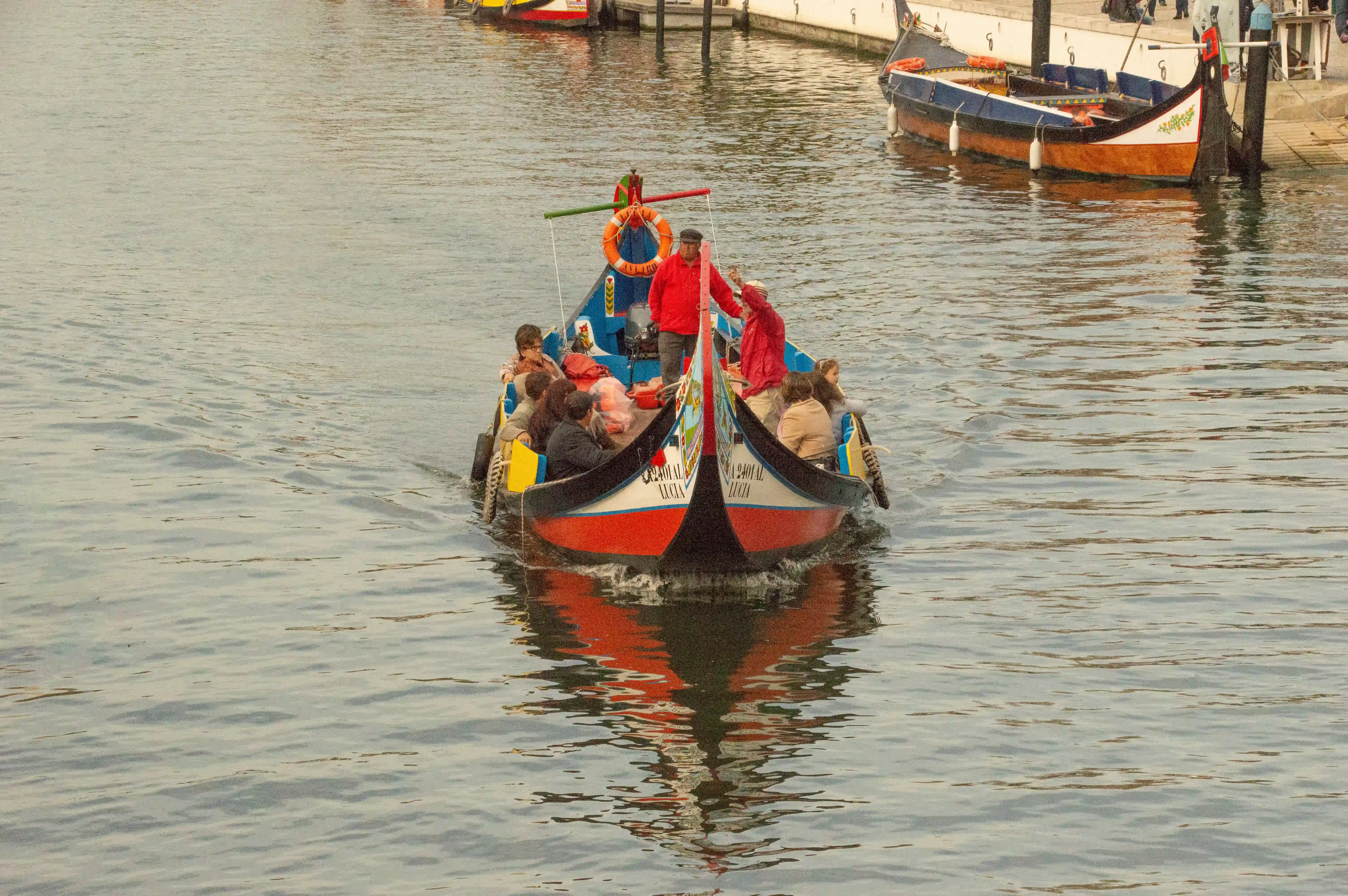 A traditional boat with passengers navigating a canal, with its reflection visible on the water's surface.