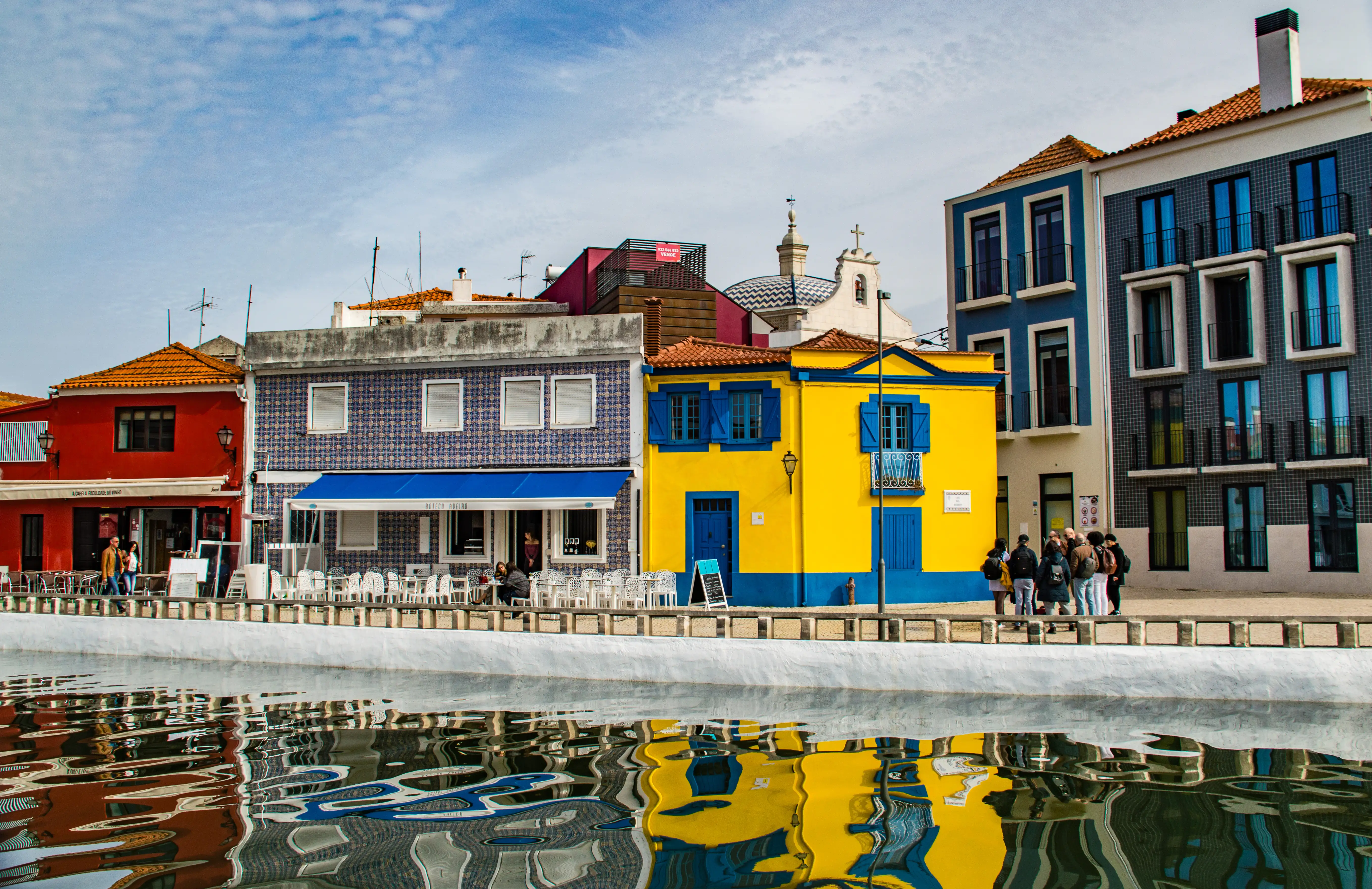 A line of brightly coloured buildings, including red, blue-tiled, yellow, blue, and grey structures, stand along a white promenade beside a calm body of water. The buildings are reflected in the water, creating abstract shapes and colours. Several people are visible on the promenade.