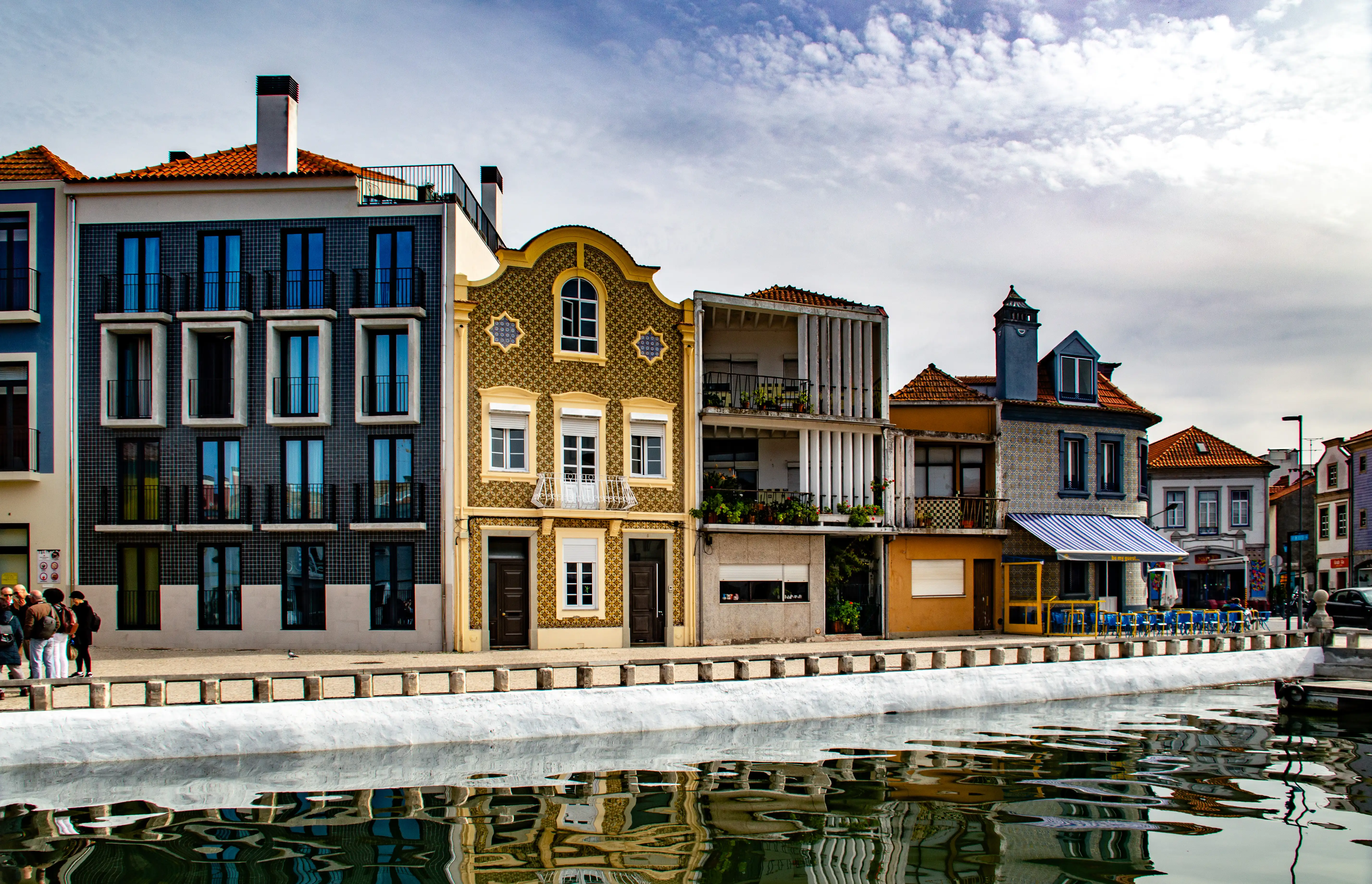  A picturesque scene showing a line of vibrant, multi-story buildings with various architectural details, including balconies and decorative facades, situated alongside a calm canal. The buildings are reflected in the water, creating a mirror image. A small group of people are visible walking along the canal's edge.
