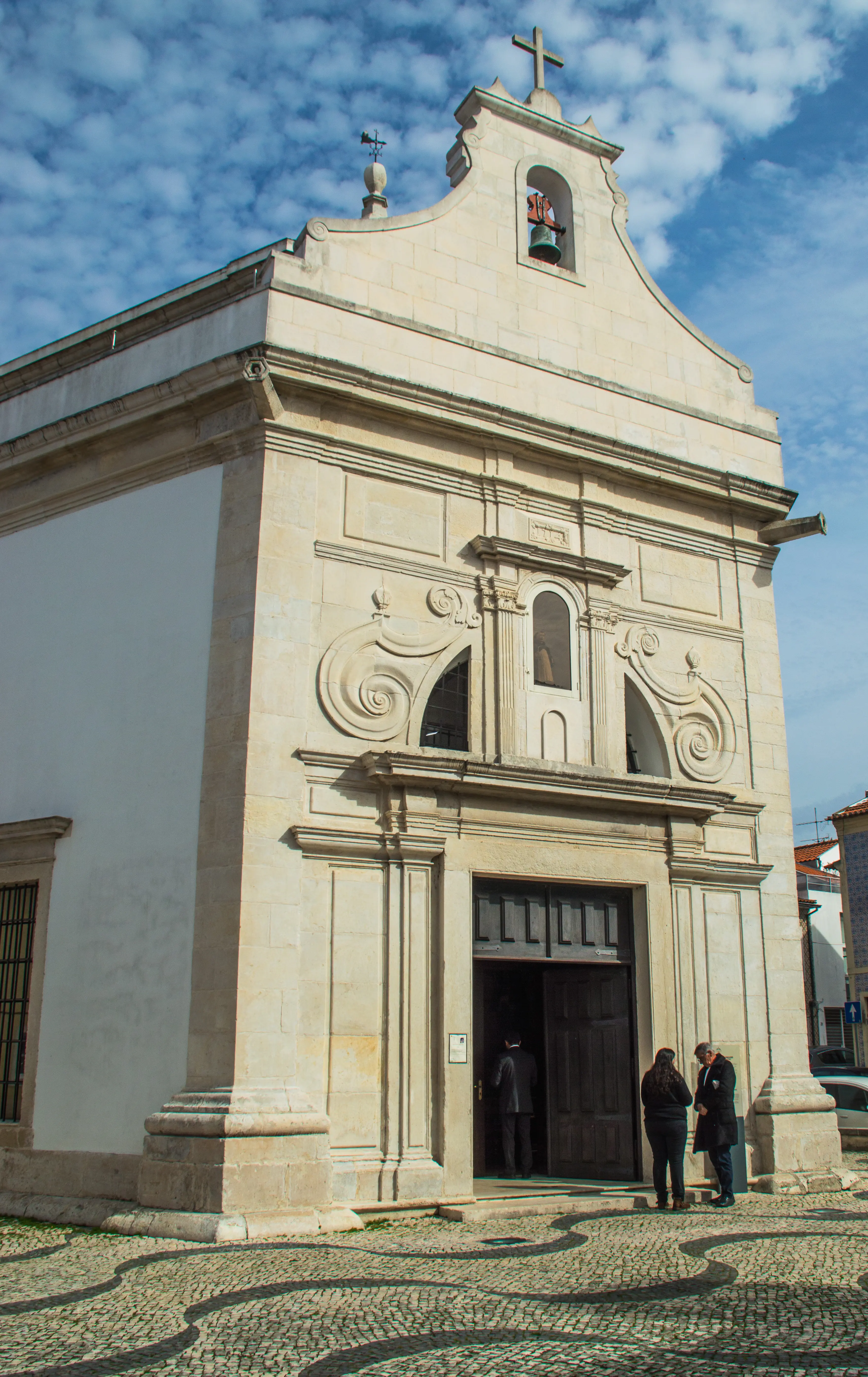 A light-coloured stone church with intricate carvings and a bell tower against a blue, cloudy sky. People are entering the church through a large wooden door.