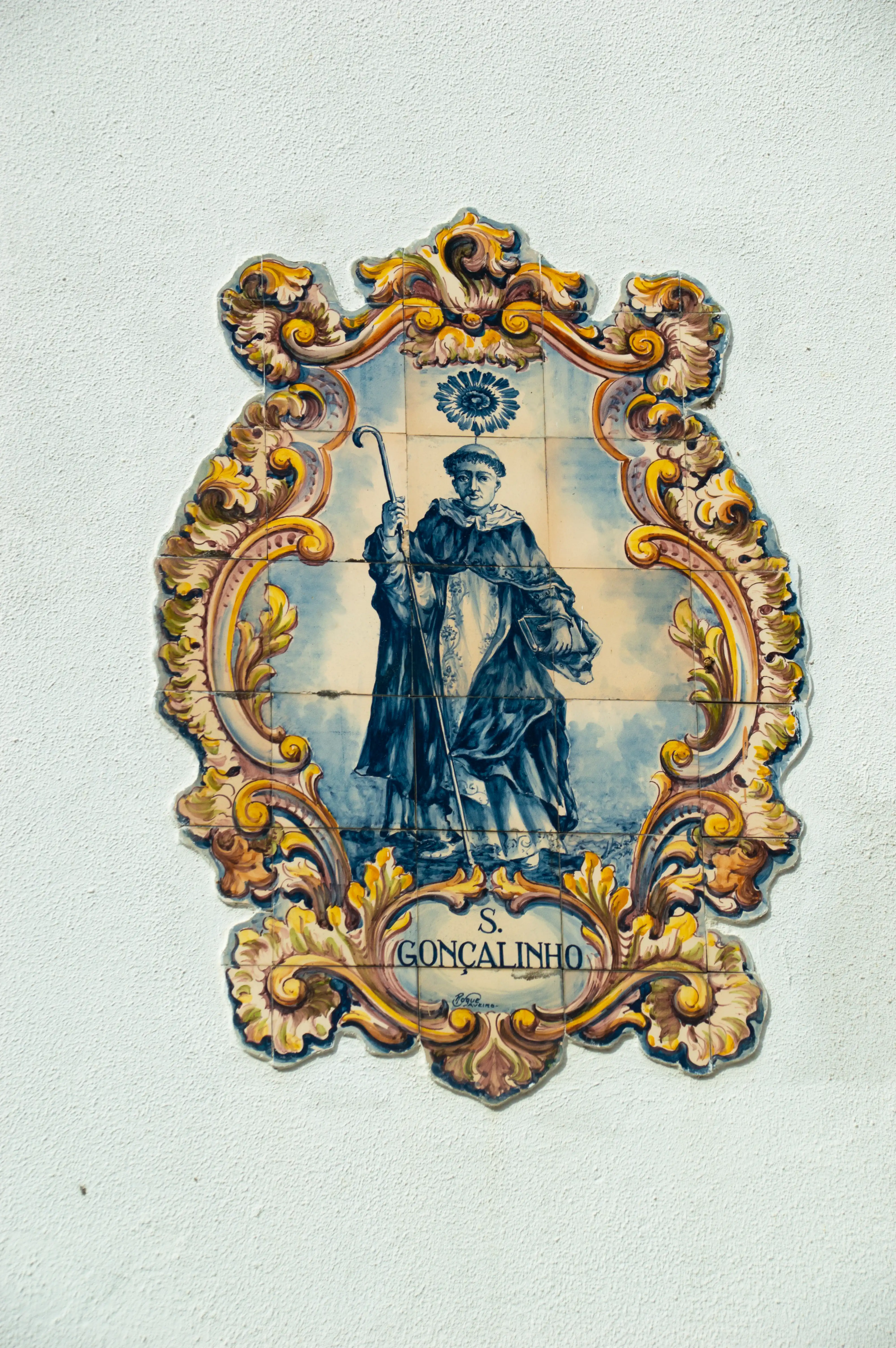 Ornate ceramic tile plaque on a white wall showing a saint in robes holding a staff, with 