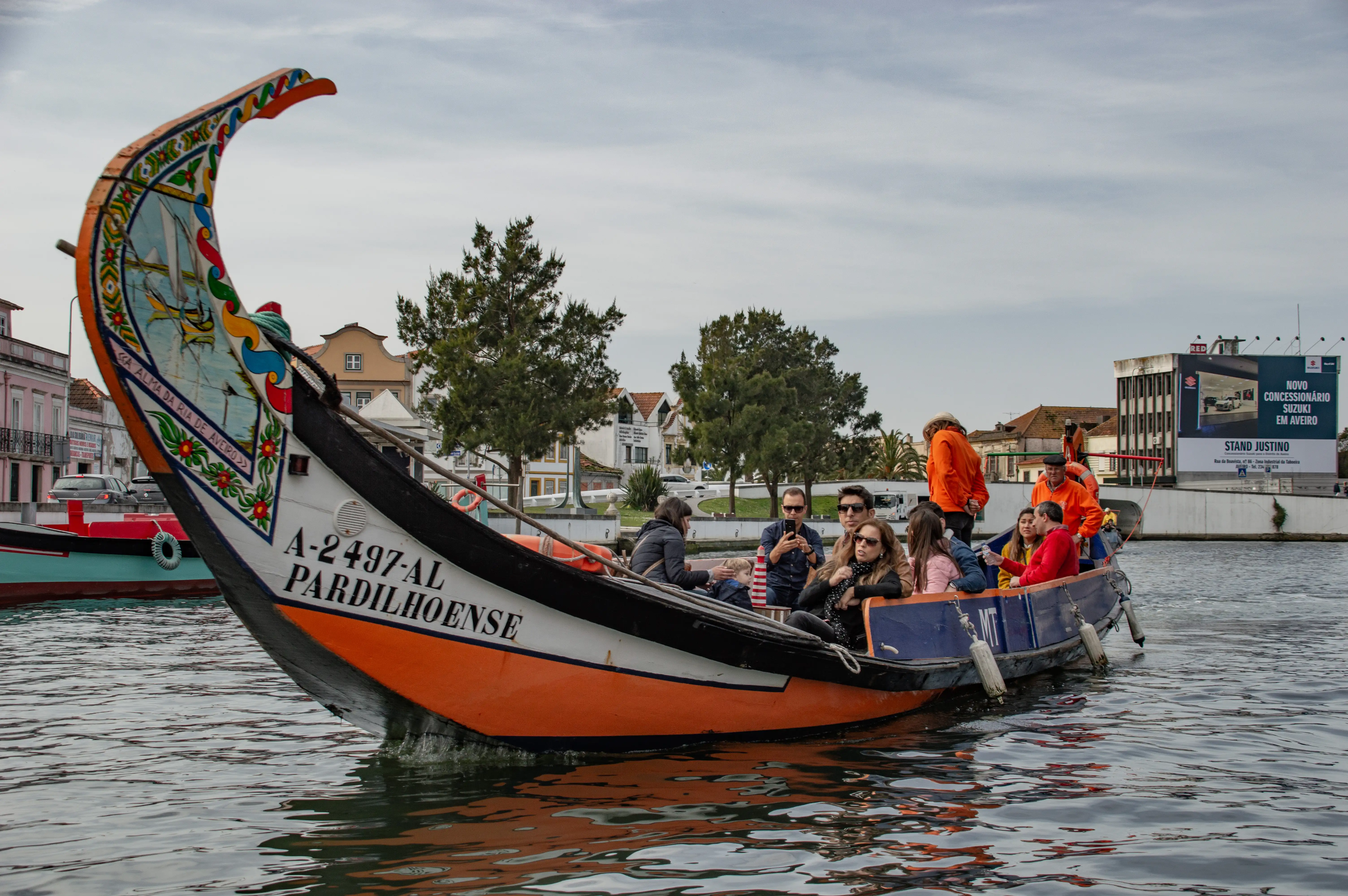 A decorated traditional Portuguese moliceiro boat carrying tourists on a canal, with buildings and trees in the background.
