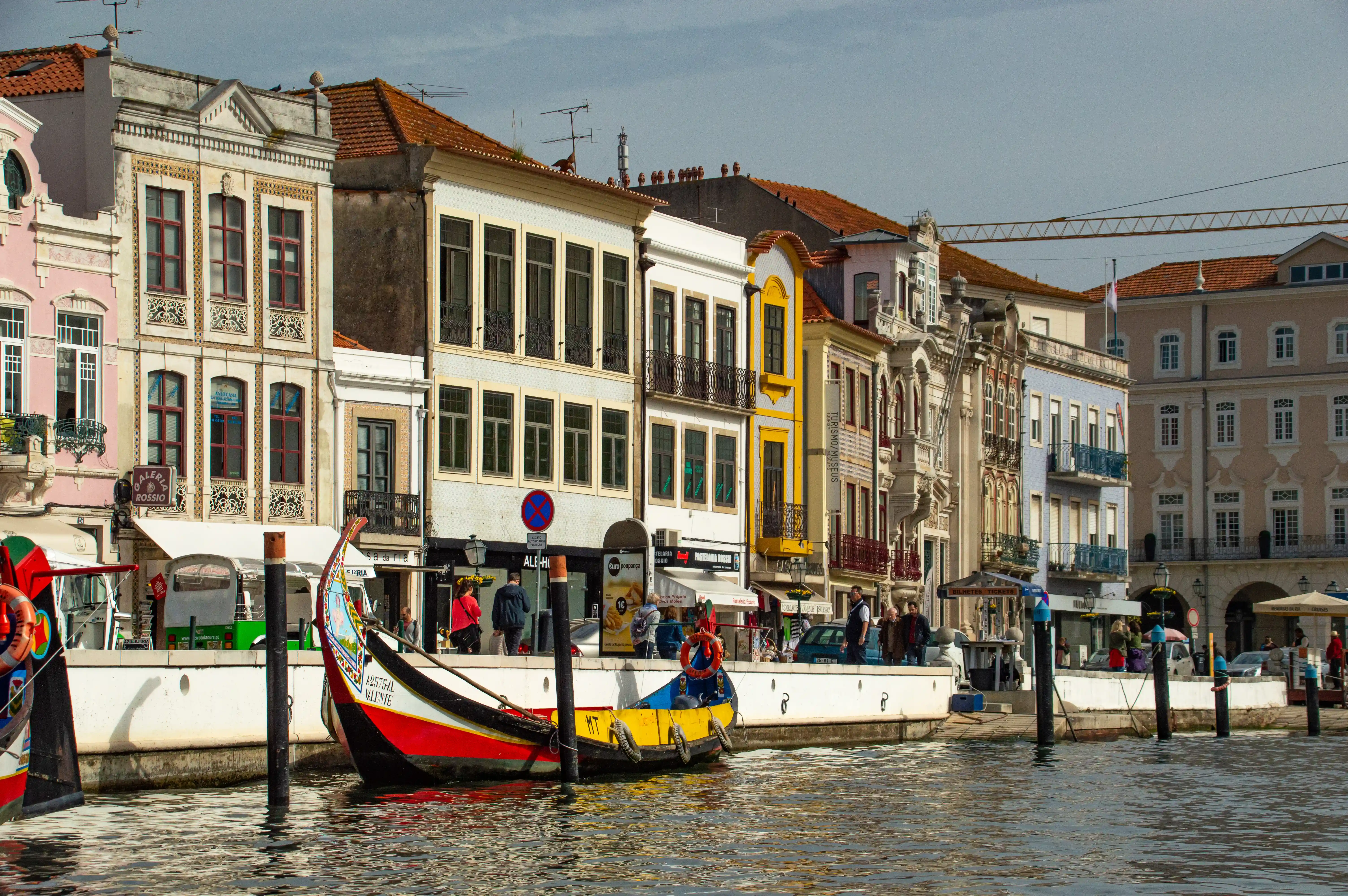 A traditional Moliceiro boat with a red, yellow, and blue hull is docked alongside a canal. In the background, a line of multi-story buildings with red tiled roofs and ornate facades in various colours such as pink, cream, yellow, and blue are visible.