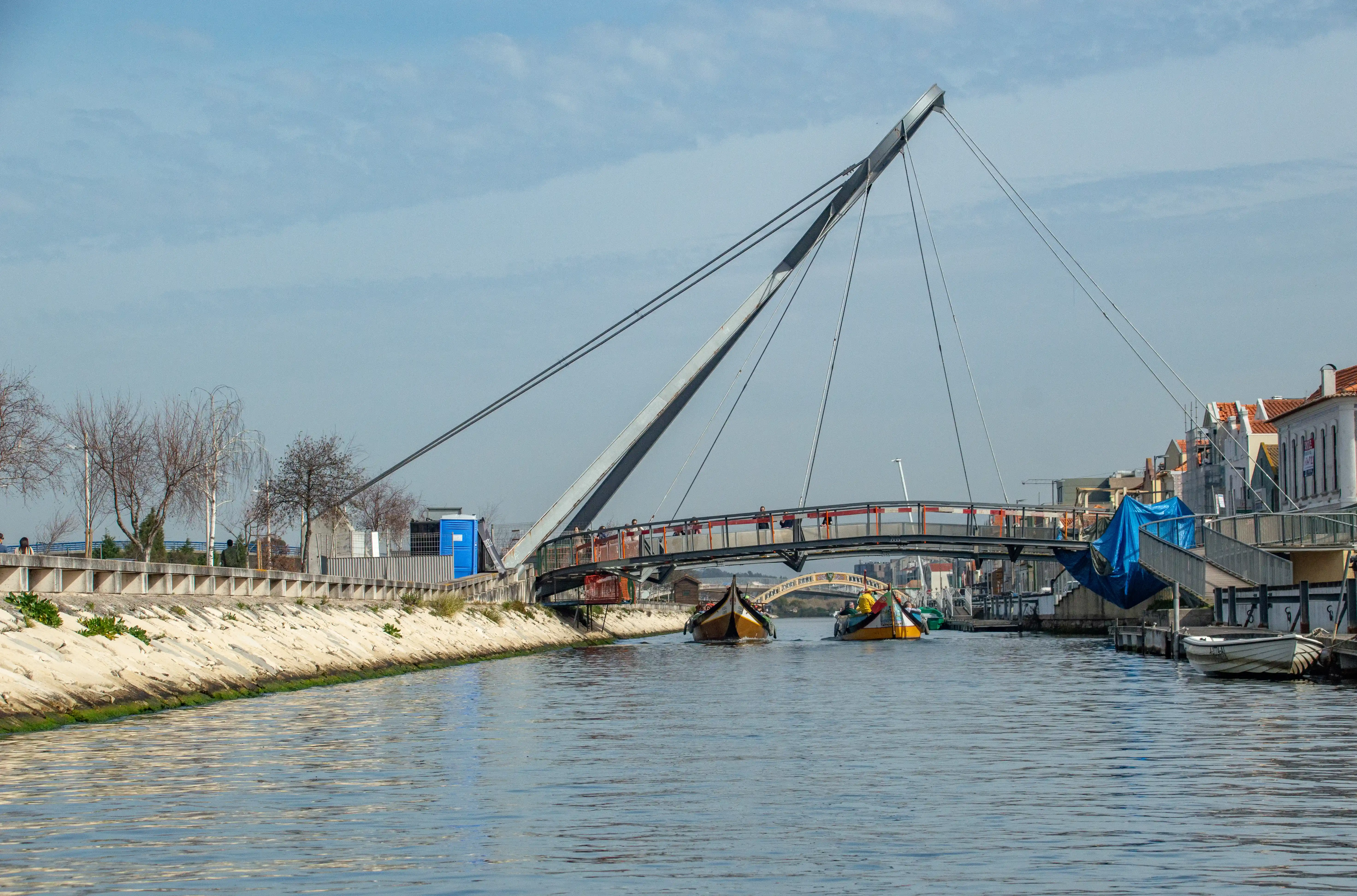 A pedestrian cable-stayed bridge with a single pylon extends over a blue canal. Two boats with distinctive colourful designs are visible on the water. The banks of the canal are lined with a concrete wall and some bare trees, with buildings and more boats visible on the right side. The sky is a pale blue with some wispy clouds.
